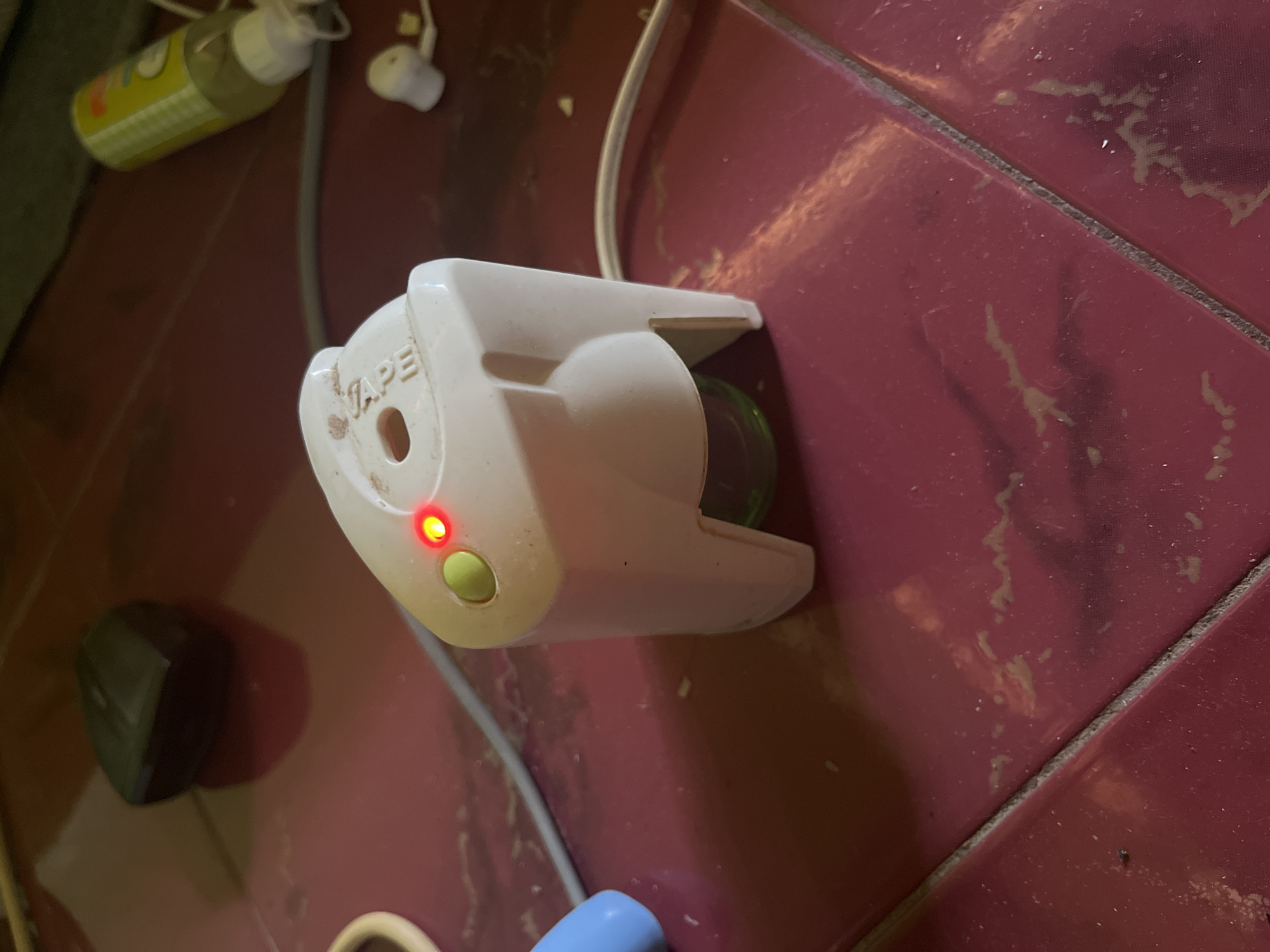 In Bumi Mulyo, Indonesia, a small, white electronic device with a green button and a red indicator light sits on a maroon tiled surface. A grey cord connects to the device and snakes across the tiles. To the left of the device, a blurry blue object is partially visible, and further left, a dark, angular object rests on the floor. In the upper right quadrant, a yellow bottle with a white cap and a faint pattern is situated near a pair of white earbuds. The tiled surface is dusty and appears worn.