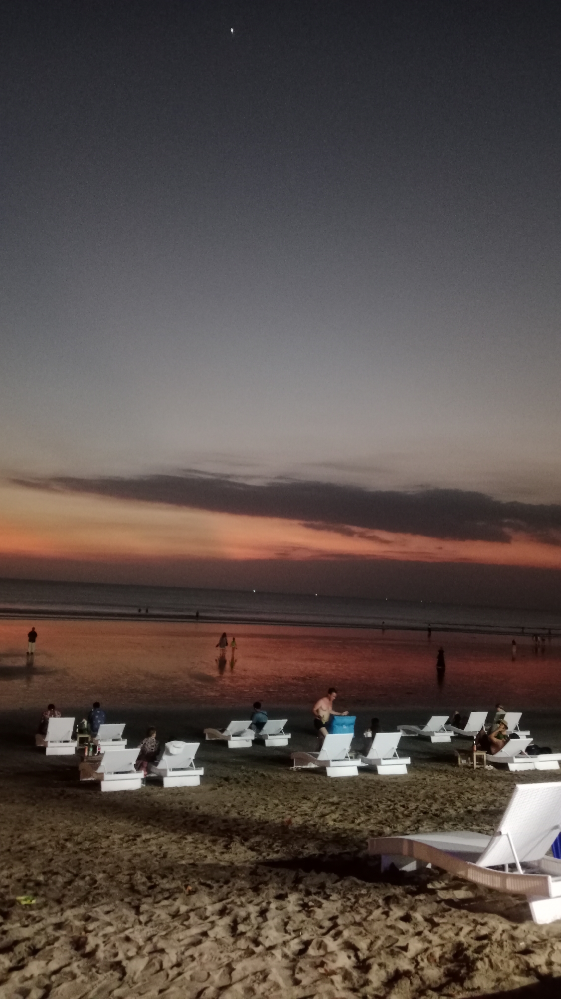 A panoramic view captures the Kuta beach in Bali, Indonesia, at dusk. The sky is a gradient of deep indigo at the top, transitioning to soft grey and then to vibrant hues of orange and pink near the horizon, indicating the setting sun. A single bright star is visible in the upper left quadrant of the sky. The ocean is calm, reflecting the colorful sky, with a gentle surf lapping at the shore.
The sandy beach in the foreground is scattered with numerous white lounge chairs. Some chairs are occupied by people, while others are empty. In the mid-ground, several individuals are walking or standing near the water's edge. The wet sand reflects the twilight sky, creating a mirror-like effect.
In the foreground, a white lounge chair is prominently positioned, empty and casting a shadow on the sand. The sand itself is textured and appears damp in some areas.
Several groups of people are seen relaxing on the lounge chairs. In the mid-ground, a man in swim trunks is standing next to a blue cooler or bag, facing away from the camera. Other individuals are seated or reclining on the lounge chairs, some appearing to engage in conversation. Further back, along the shoreline, a few people are visible walking or wading in the shallow water. The overall atmosphere is serene and relaxed, typical of a beach at sunset.