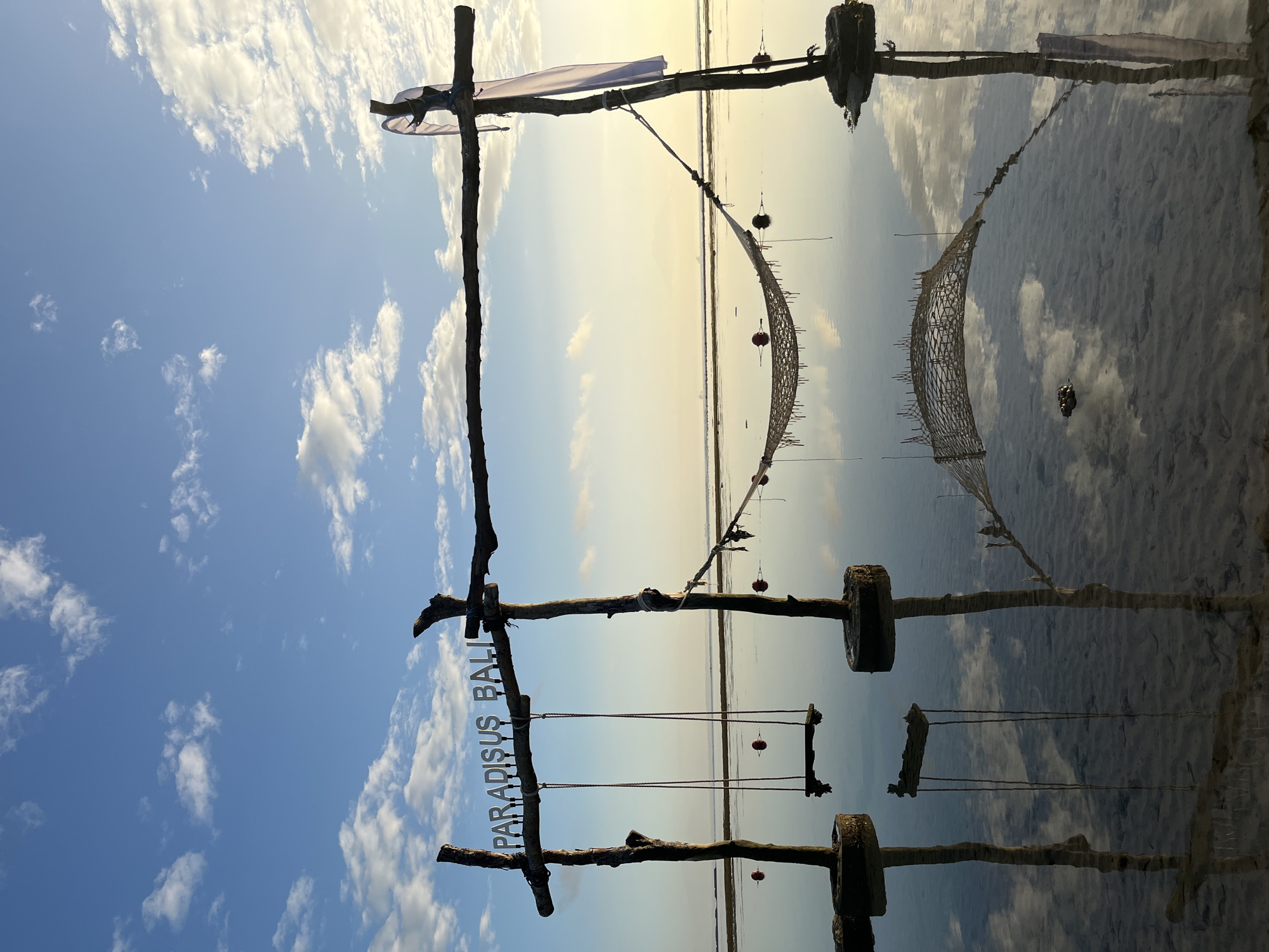 This is a serene, low-angle, landscape photograph captured at dawn or dusk, likely at the beach in Kuta Selatan, Indonesia. The scene is dominated by a wooden structure supporting a swing and a hammock, which are reflected in the still, shallow water. The words "PARADISUS BALI" are mounted on the horizontal beam of the structure. Two red buoys are visible in the water, one near the swing and another near the hammock. The sky above is a soft blue, adorned with scattered white clouds. The water's surface acts as a mirror, reflecting the sky and the wooden structure with remarkable clarity. The overall impression is one of tranquility and natural beauty.
