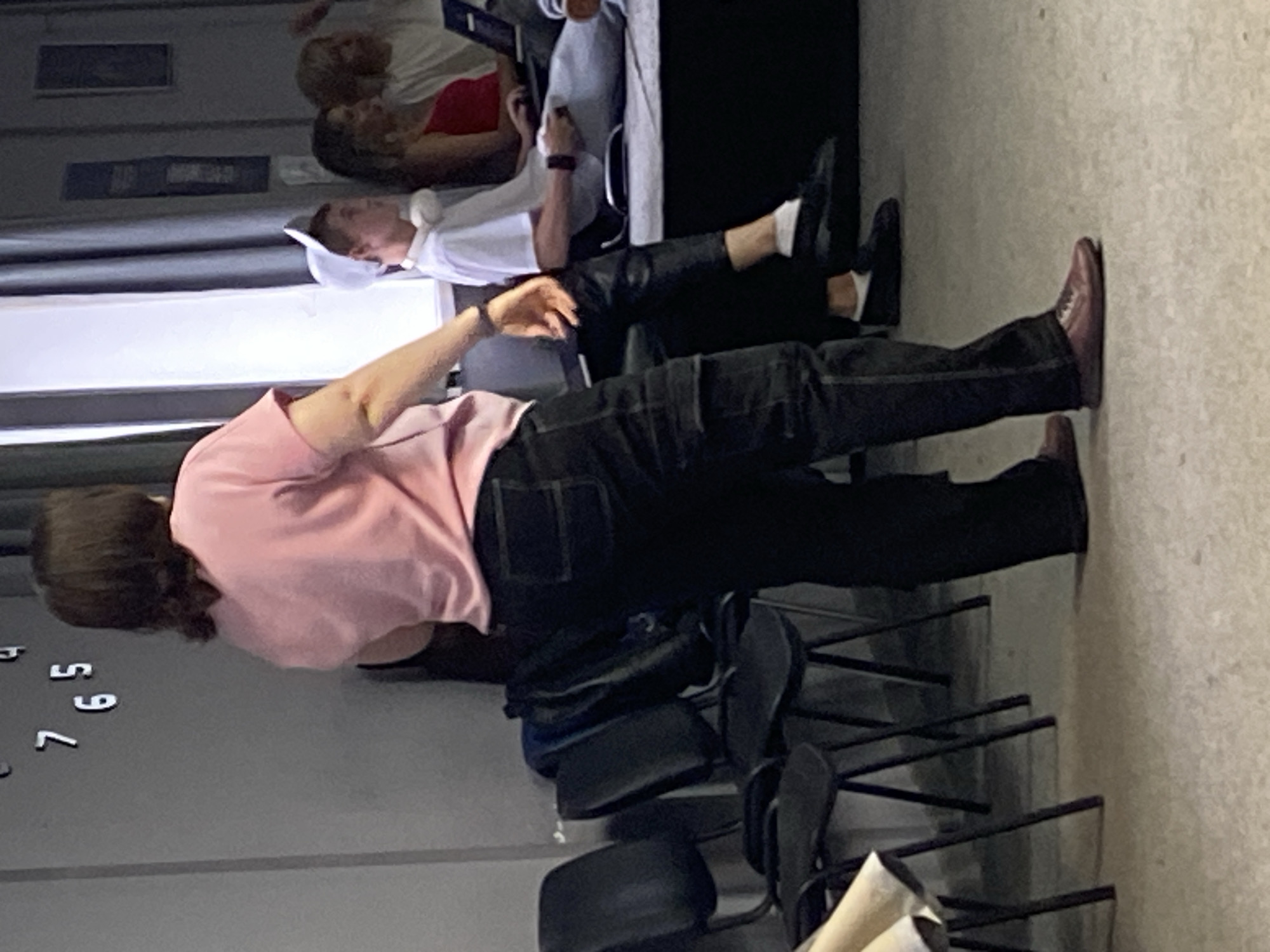 In an indoor setting, a person wearing a pink t-shirt and black pants stands facing away from the camera. Their hair is dark brown and cut to shoulder length. To the right of this individual, several other people are seated. One person, wearing a white baseball cap and headphones, is looking to the left. Another person in a red top is partially visible, and further back, a person in a white shirt is seated. There is a laptop on a surface in front of the seated individuals. The environment includes a grey wall with a clock, black chairs, and a curtained window. The floor is a light grey textured surface.