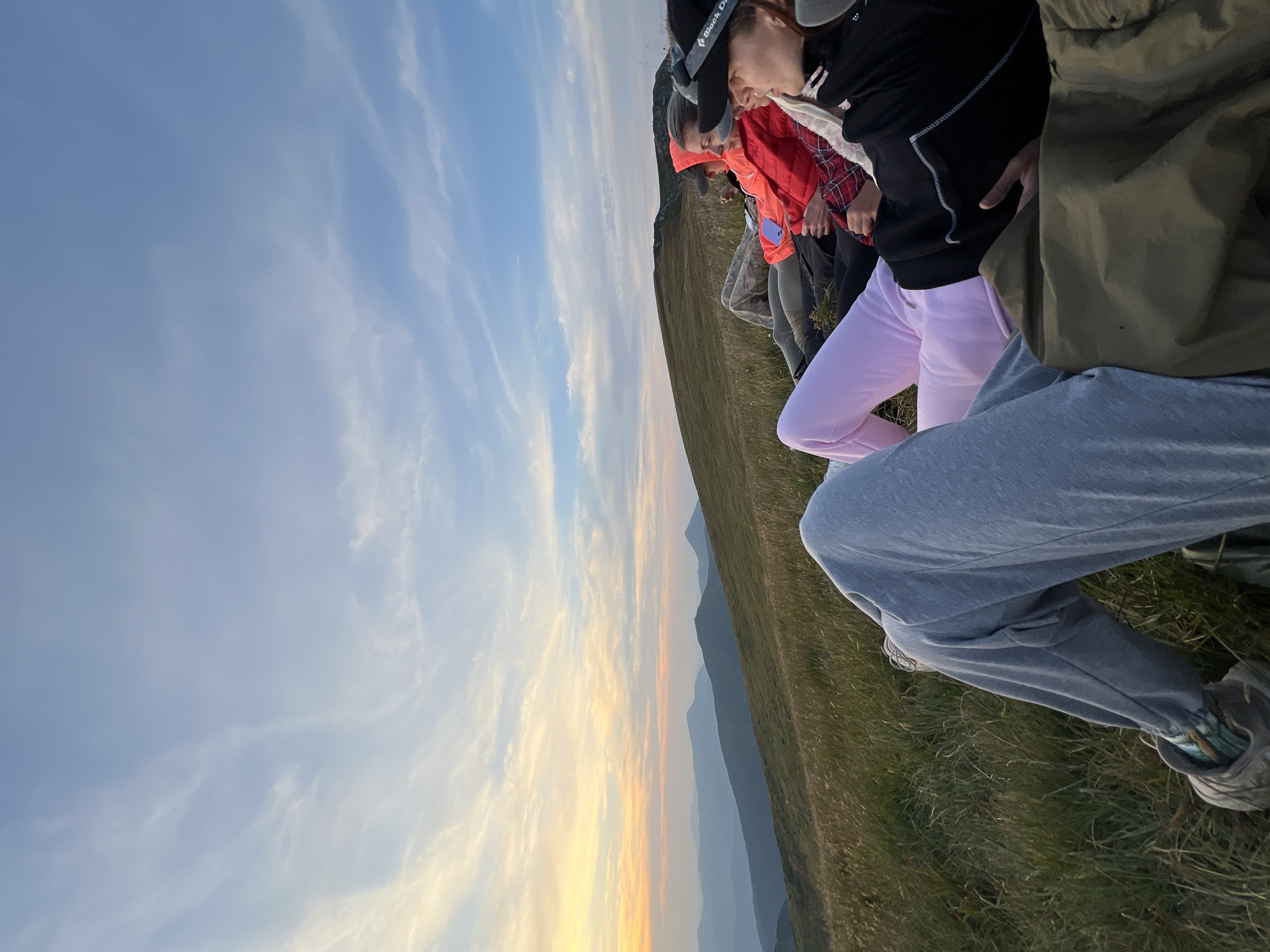 Four individuals are observed from a low-angle perspective, reclining on a grassy mountainside, likely in the vicinity of Hoverla, Ukraine, during either sunrise or sunset, evidenced by the warm, pastel hues illuminating the sky and distant mountain ranges. The individuals are casually dressed in outdoor attire, with one person in the foreground wearing grey sweatpants and hiking shoes. To their right, another person is clad in bright pink sweatpants and a white top, while a third wears a red puffer jacket and a fourth is partially visible in the background. The environment is characterized by rolling green hills covered in tall, dry grass, with a clear sky featuring wispy clouds. The overall atmosphere suggests a moment of shared contemplation or rest amidst a scenic natural landscape.