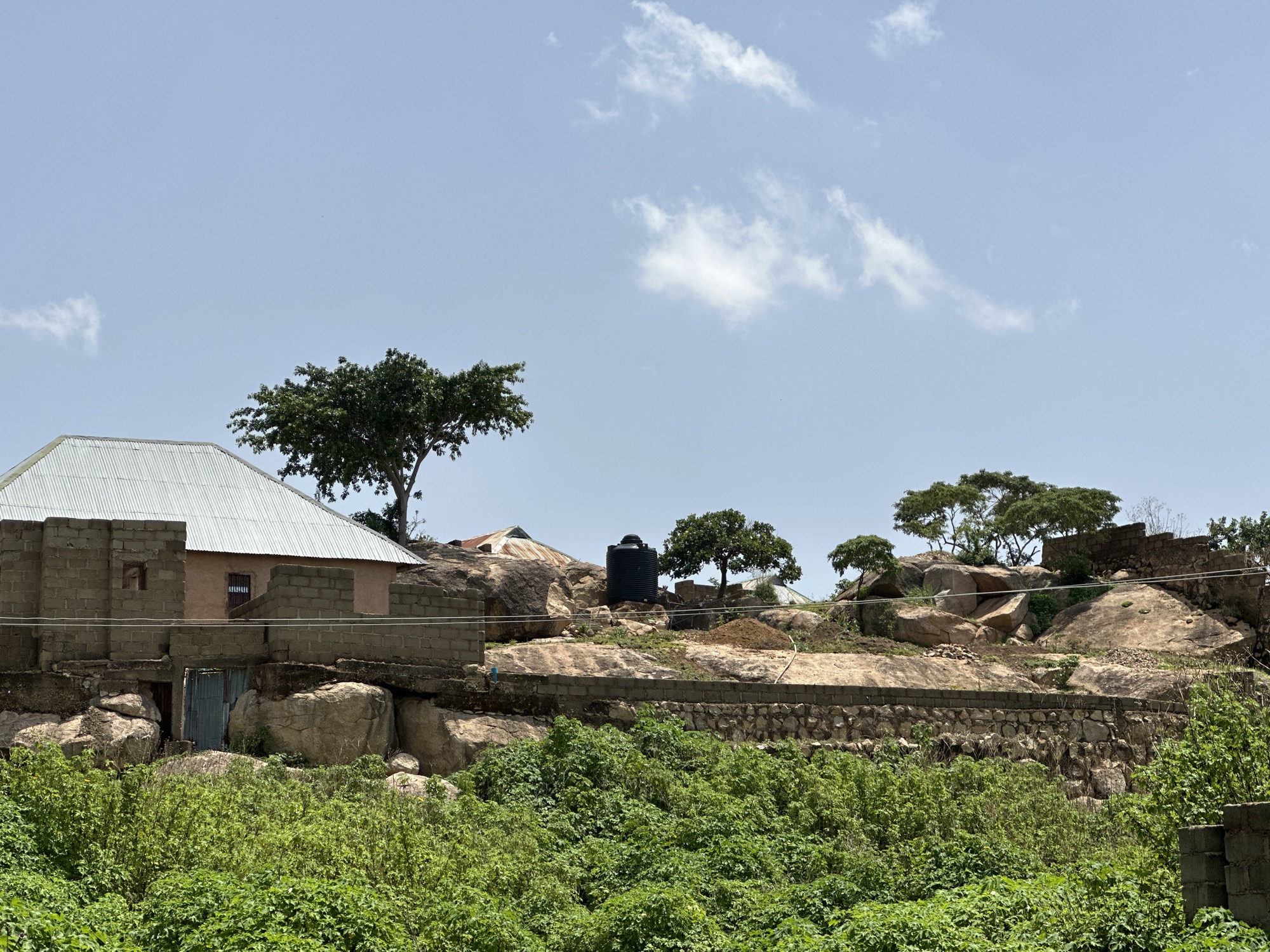 The image depicts a rural scene in Jos, Nigeria, characterized by a rocky terrain dotted with vegetation and several buildings. In the foreground, lush green bushes dominate, suggesting a fertile environment. Behind the bushes, a stone wall separates the lower area from a higher rocky elevation.
On this elevated ground, a prominent building with a corrugated metal roof and brick walls stands out. Adjacent to it, other structures with similar roofing materials are visible, some partially obscured by trees. A large black water tank is situated on the rocks, indicating a need for water storage. The landscape is characterized by large, weathered boulders and sparse vegetation, with several trees adding greenery to the scene. The sky is a pale blue with scattered white clouds, suggesting fair weather. The overall impression is one of a settlement integrated with the natural rocky landscape of Jos.