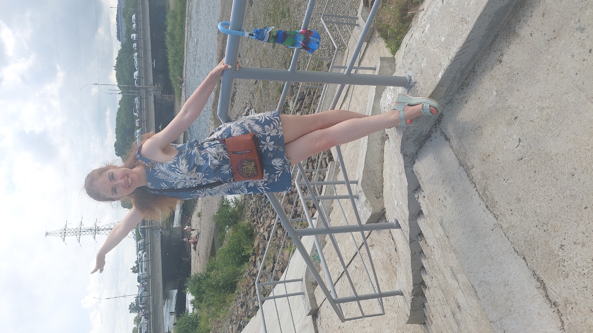 A young Caucasian woman with long, auburn hair is posing on concrete stairs next to a metal railing. She is wearing a blue floral print dress, a tan crossbody bag with a lotus flower emblem, and light blue wedge sandals. Her left arm is extended upwards and to the left, while her right hand rests on the railing. Behind her, a river is visible, with a bridge crossing it in the distance. Cars are lined up on the bridge, and a large power line tower stands tall against a cloudy sky. The woman is smiling at the camera. The scene appears to be set in Komsomolsk-on-Amur, Russia, likely near the Amur River embankment.