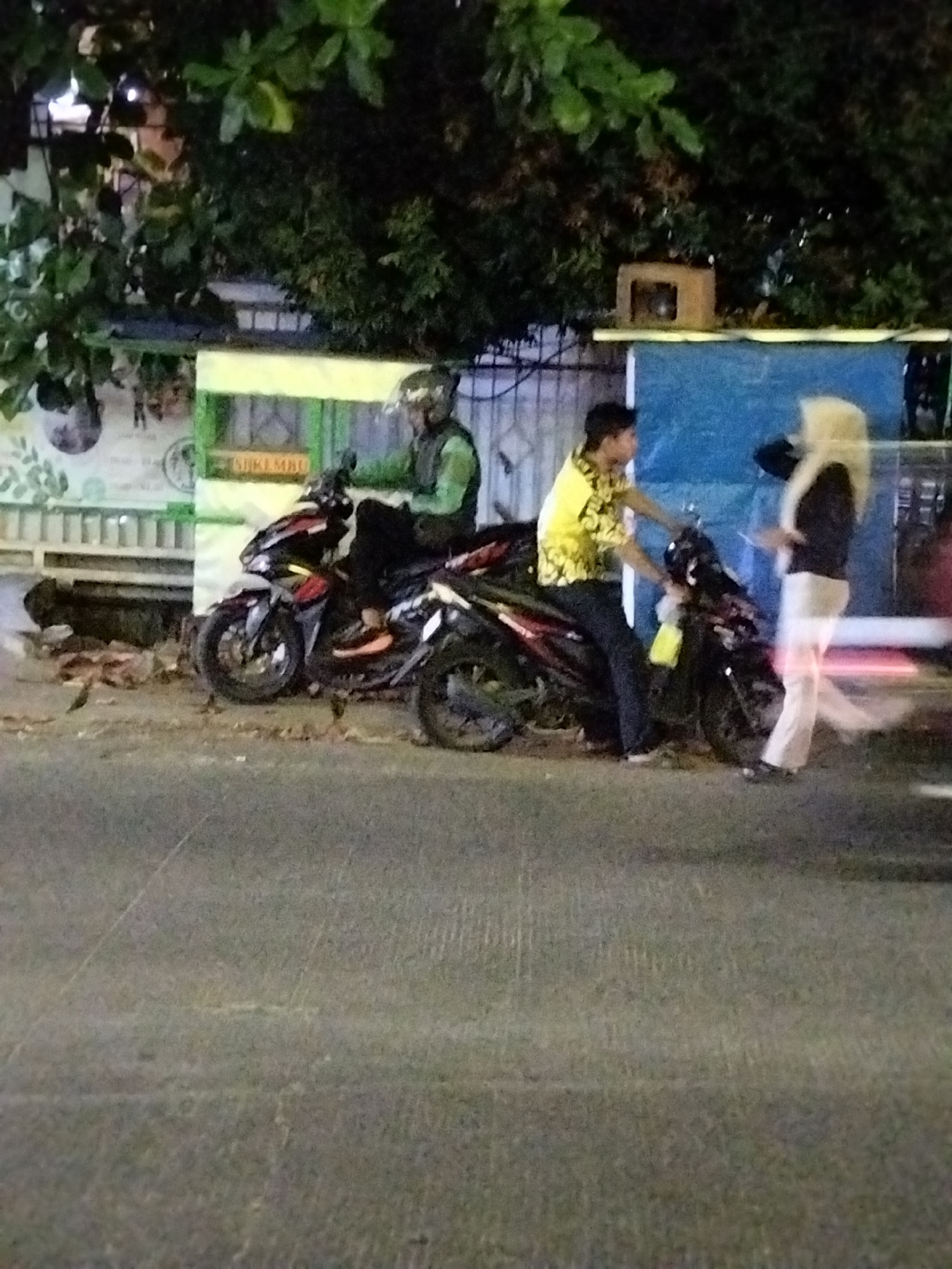 The image captures an outdoor scene at night, likely on a street in South Tangerang, Indonesia, given the presence of palm trees and the architectural style of the buildings. In the foreground, a paved road is visible. Behind the road, there are two motorcycles parked. The first motorcycle is red and black, with a rider wearing a green jacket and a helmet. The second motorcycle is black, with a rider in a bright yellow, patterned shirt and dark pants. This rider appears to be interacting with a person standing next to the motorcycle, who is wearing a light-colored hijab and dark clothing. The background features a low-rise building with a blue facade and some signage. Green foliage, possibly from trees, hangs over the scene. The lighting suggests artificial illumination from streetlights or nearby buildings, creating a nighttime atmosphere.