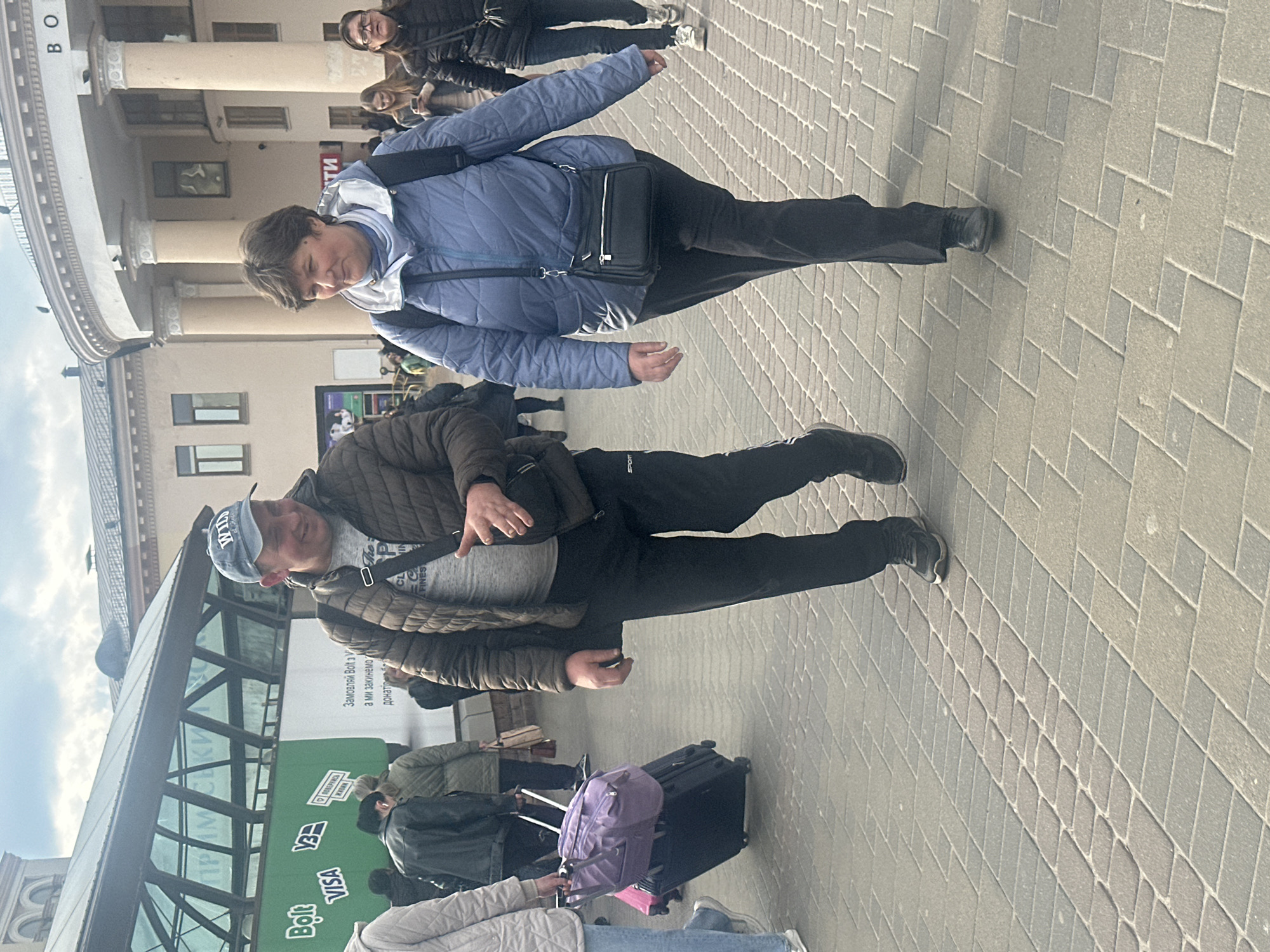 The image captures an outdoor scene in Kyiv, Ukraine, likely a pedestrian area or public square in front of a large building. The time of day is daylight, with sunlight suggesting a clear or partly cloudy sky.
In the foreground, a man is visible wearing a brown quilted jacket, a grey t-shirt, black athletic pants with "SPORT" visible on the leg, and a light-colored baseball cap with "WILD" printed on it. He has a black shoulder bag. He is looking downwards, with his right hand holding a small, dark object. Slightly behind him, a woman walks in a blue quilted jacket over a white hoodie, black pants, and a black cross-body bag. She also appears to be looking down or to her side.
In the mid-ground and background, other individuals are present, some carrying luggage, including a purple backpack and a black wheeled suitcase, suggesting travel or transit. The setting includes patterned grey and lighter grey paving stones. To the left, a modern glass-covered structure or walkway is visible, reflecting the sky. In the upper background, a grand, classic-style building with multiple columns stands, with "BO" visible as part of its signage.
Several signs and text are visible: "Bolt," "VISA," and a larger green sign with Ukrainian text that reads "Замовляй Bolt з V а ми закинемо донаті" (Order Bolt with V and we will donate), indicating the use of Bolt services for charity. The overall atmosphere is that of a busy urban environment with people on the move.