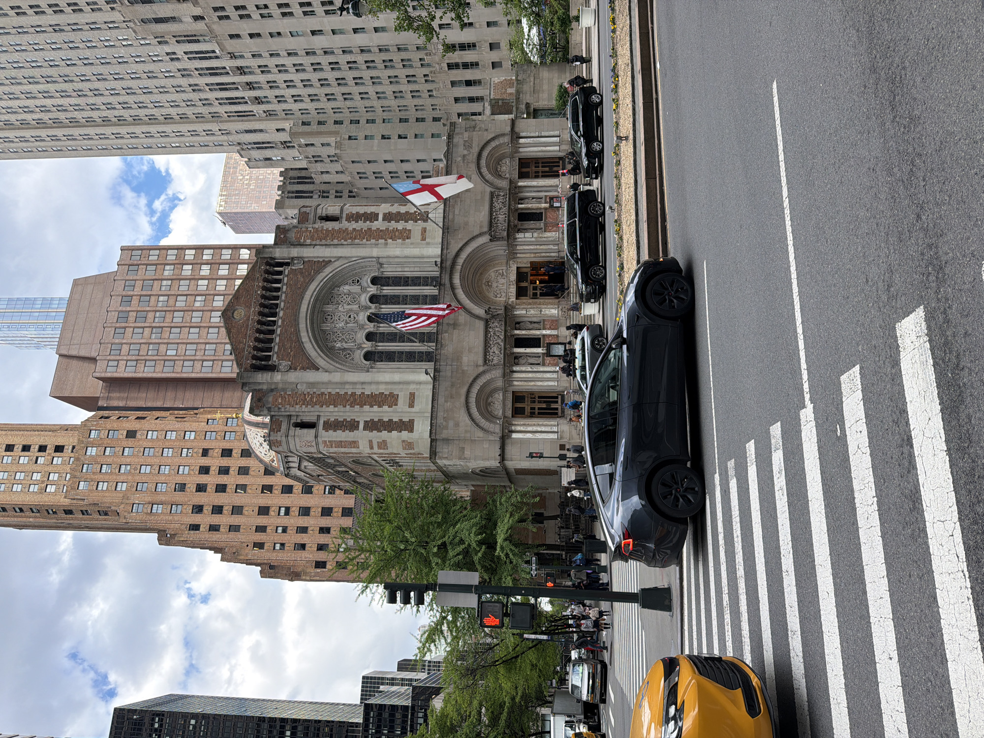 The image captures a street scene in New York City, focusing on the Stephen Wise Free Synagogue on West 68th Street. The synagogue is adorned with an American flag and another flag bearing a red cross. Several black cars are parked along the side of the street in front of the synagogue. A traffic light displays a red hand. Pedestrians wait at the crosswalk. A yellow taxi is also visible. Tall buildings dominate the background, contributing to the urban setting.
