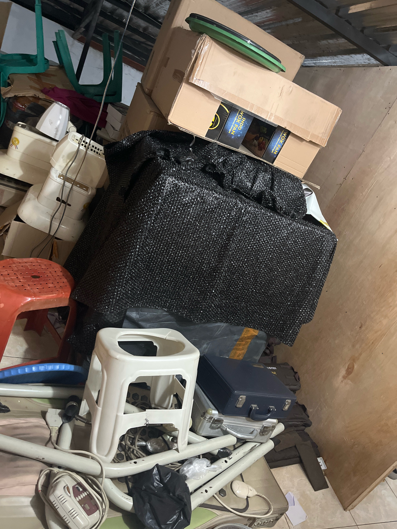 The image depicts a cluttered indoor space, possibly a storage area or a room in disarray, located in Matesih, Indonesia. The scene is dominated by stacked cardboard boxes, some open and revealing contents like what appear to be cooking pans with packaging. A large, black, textured object, possibly a tarpaulin or a woven covering, is prominently placed in the center.
Several household appliances, including what seem to be parts of blenders or similar devices, are visible. There are also various seating items, such as a bright red plastic stool and a white plastic stool. Other objects include a dark blue briefcase, a silver suitcase, and what appears to be part of a medical support device or frame. Electrical cords and wires are scattered throughout the area.
The environment shows signs of disorganization, with items piled haphazardly. The background is a wooden wall, suggesting an interior setting. The lighting is dim, contributing to a cluttered and somewhat dingy atmosphere. No individuals are present in the frame.
