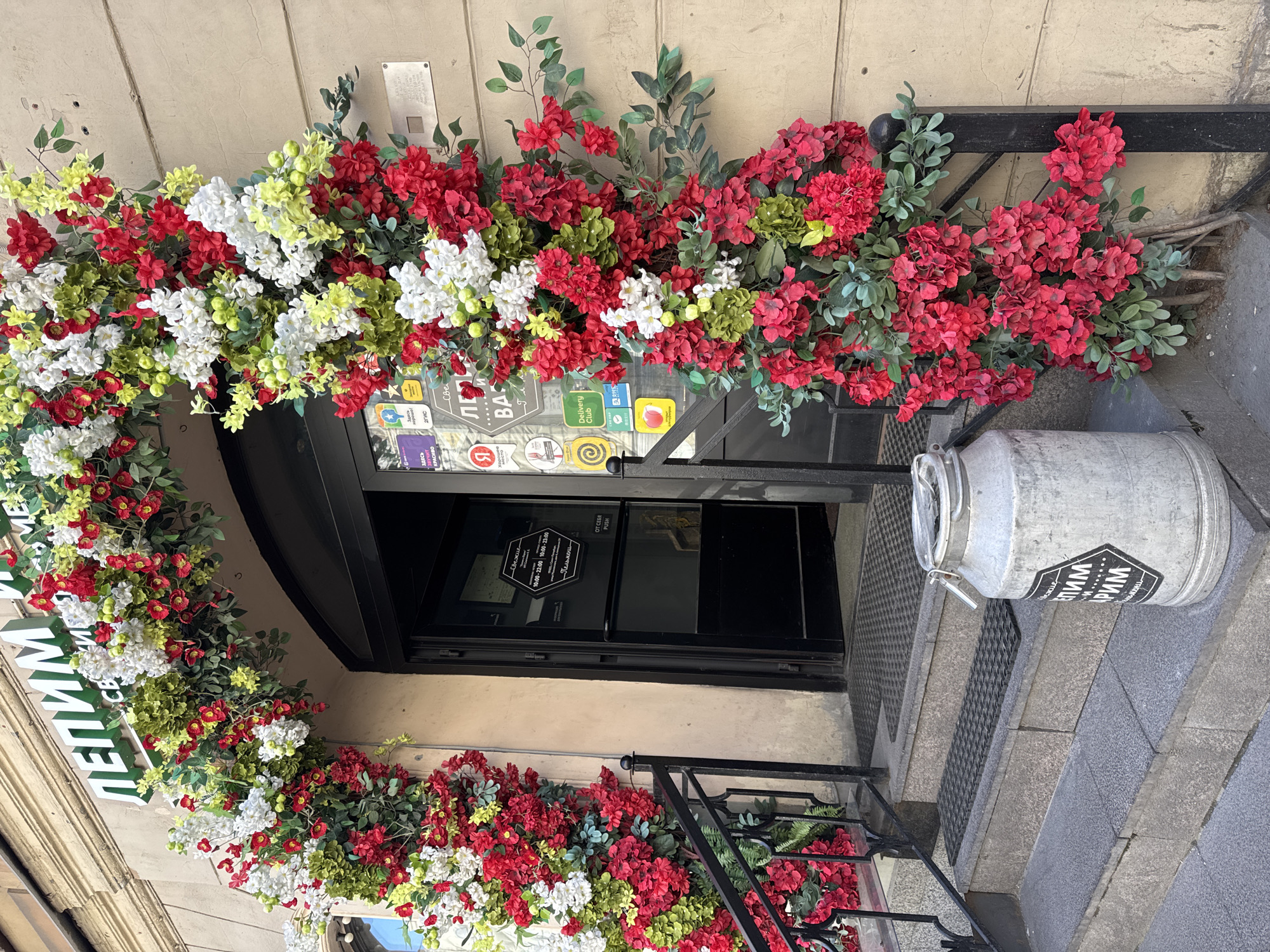The image shows the entrance to a shop or cafe adorned with a lush floral archway. The arch is composed of numerous artificial flowers in shades of red, white, and green, interspersed with faux foliage. Some of the flowers appear to be hydrangeas, while others resemble smaller blossoms.
The entrance itself is a dark-colored double door, with one side slightly ajar, revealing a glimpse of the interior. Several stickers are affixed to the glass of the door, including what appear to be delivery service logos and promotional decals. One sticker prominently displays the text "Delivery Club". Above the doorway, signage in green letters on a white background reads "ЛЕПИМ И ВЫПЕКАЕМ", which translates to "We Mold and Bake". This suggests the establishment is a bakery or a place that serves baked goods.
To the left of the entrance, a black metal railing leads up a set of concrete stairs. To the right of the stairs, standing on a lower step, is a large, distressed metallic milk can with a lid and handle. A dark hexagonal sticker with white text and a border is attached to the front of the can, and the text is partially legible as "ЛЕПИМ И ВЫПЕКАЕМ".
The building's facade is a light beige color with visible texture. The overall scene suggests a welcoming and decorative storefront, likely in a pedestrian area of the city, given the presence of delivery service stickers. Based on the Cyrillic signage and the architectural style, the location is Saint Petersburg, Russia. No people are visible in the image. The lighting suggests it is daytime, with natural light illuminating the scene.