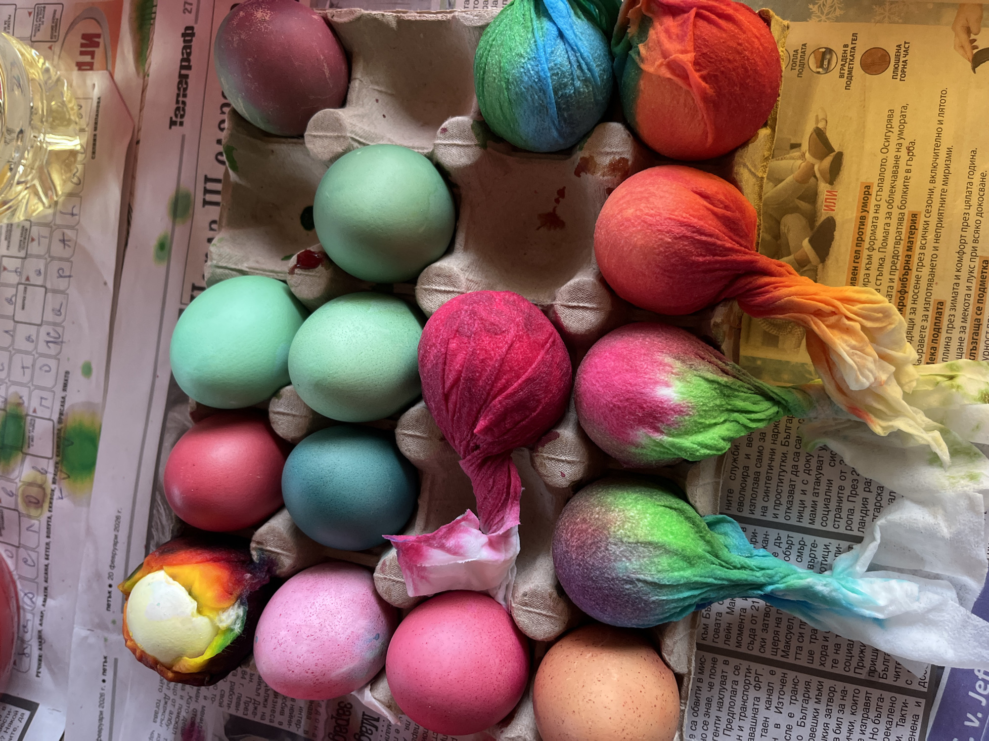 The image depicts a cardboard egg carton filled with dyed Easter eggs, resting on a newspaper. The eggs display a variety of colors including red, green, blue, orange, pink, purple, and brown, with some showing a tie-dye effect achieved with cloth wraps that are still partially attached. One egg in the bottom left corner has been partially peeled, revealing a yellow-white interior. The newspaper beneath the egg carton contains Bulgarian text, and the newspaper date, "20 февруари 2020 г.", translates to February 20, 2020. The location in Kranevo, Bulgaria is not discernible from the image alone.