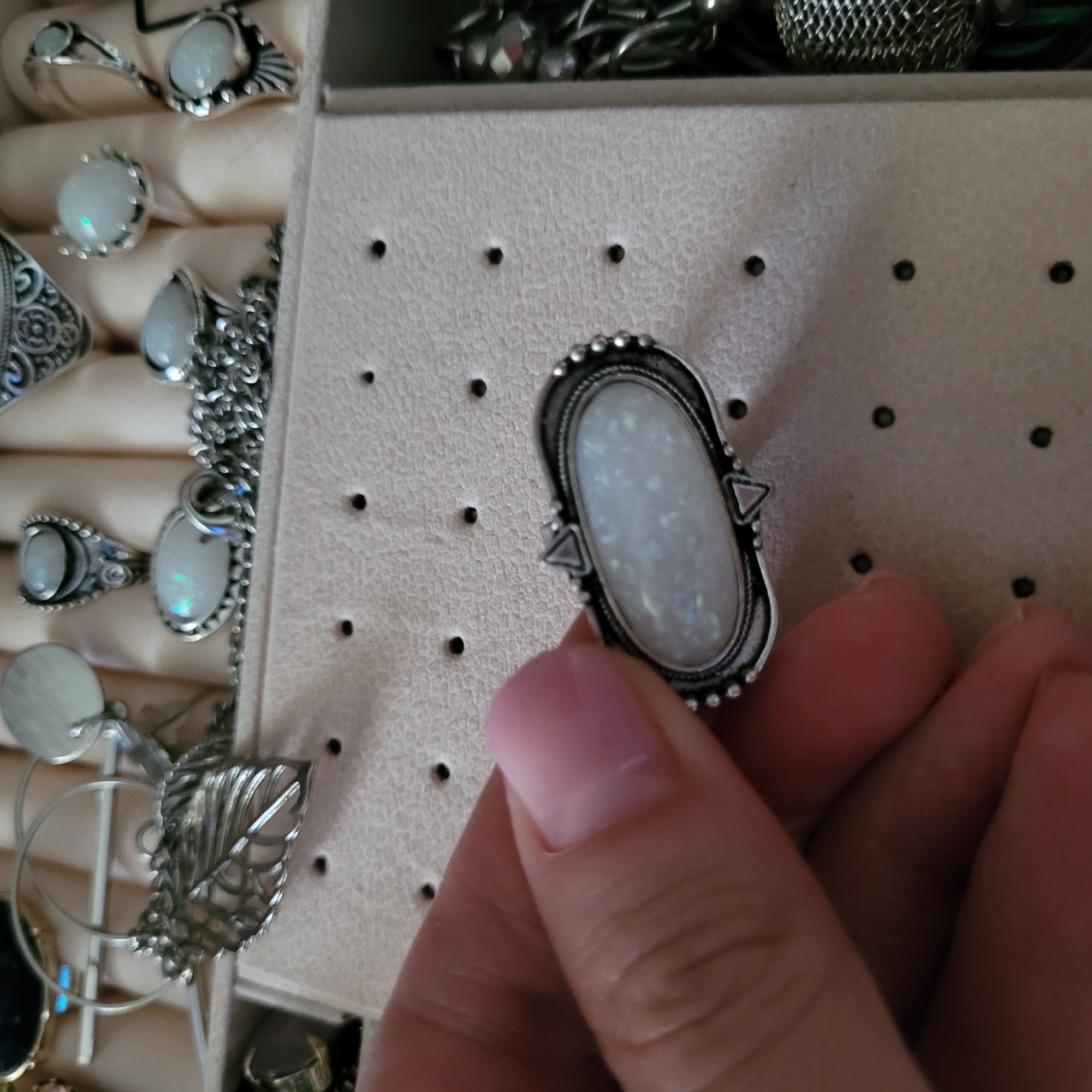 A close-up, eye-level shot shows a hand holding an oval-shaped ring against the backdrop of a jewelry organizer. The hand, with a pale pink fingernail on the index finger, grasps the ring, which features a prominent, iridescent white stone, likely an opal, set in an ornate silver-toned metal band. The ring's bezel is adorned with small metallic beads, and two triangular embellishments flank the stone on its shorter sides.
The background reveals a light beige or cream-colored jewelry organizer made of a soft, textured material, possibly felt or velvet. It has multiple rows of small, dark holes, suggesting it is designed to hold earrings or studs. Other rings are visible in the organizer, mostly with similar light-colored, possibly opal, stones and silver-toned settings. Some rings feature simple solitaire designs, while others display more intricate metalwork, including a distinctive leaf-shaped piece. Behind the organizer, out of focus, are glimpses of additional metallic objects, suggesting more jewelry storage. The scene takes place indoors under good lighting. No text is visible in the image.