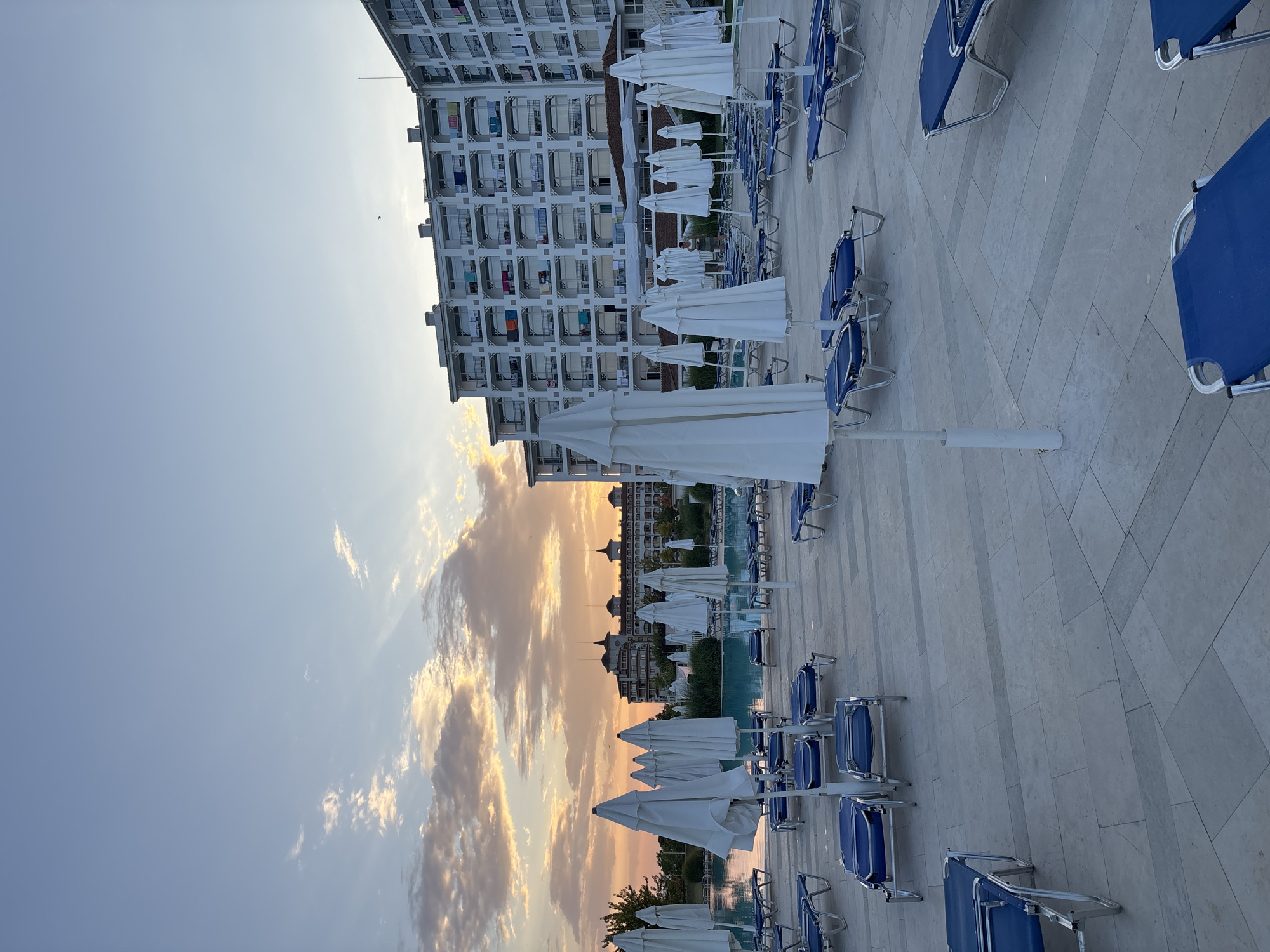 The image depicts a hotel pool area in Byala, Bulgaria, at sunset. Numerous closed white beach umbrellas and blue lounge chairs are arranged on a paved patio surrounding a swimming pool. In the background, a large, multi-story white hotel building with balconies is visible, and beyond that, another building with a distinctive architectural style. The sky is filled with dramatic clouds illuminated by the setting sun, casting warm hues of orange and yellow. The scene is tranquil, suggesting the end of a day at a resort.