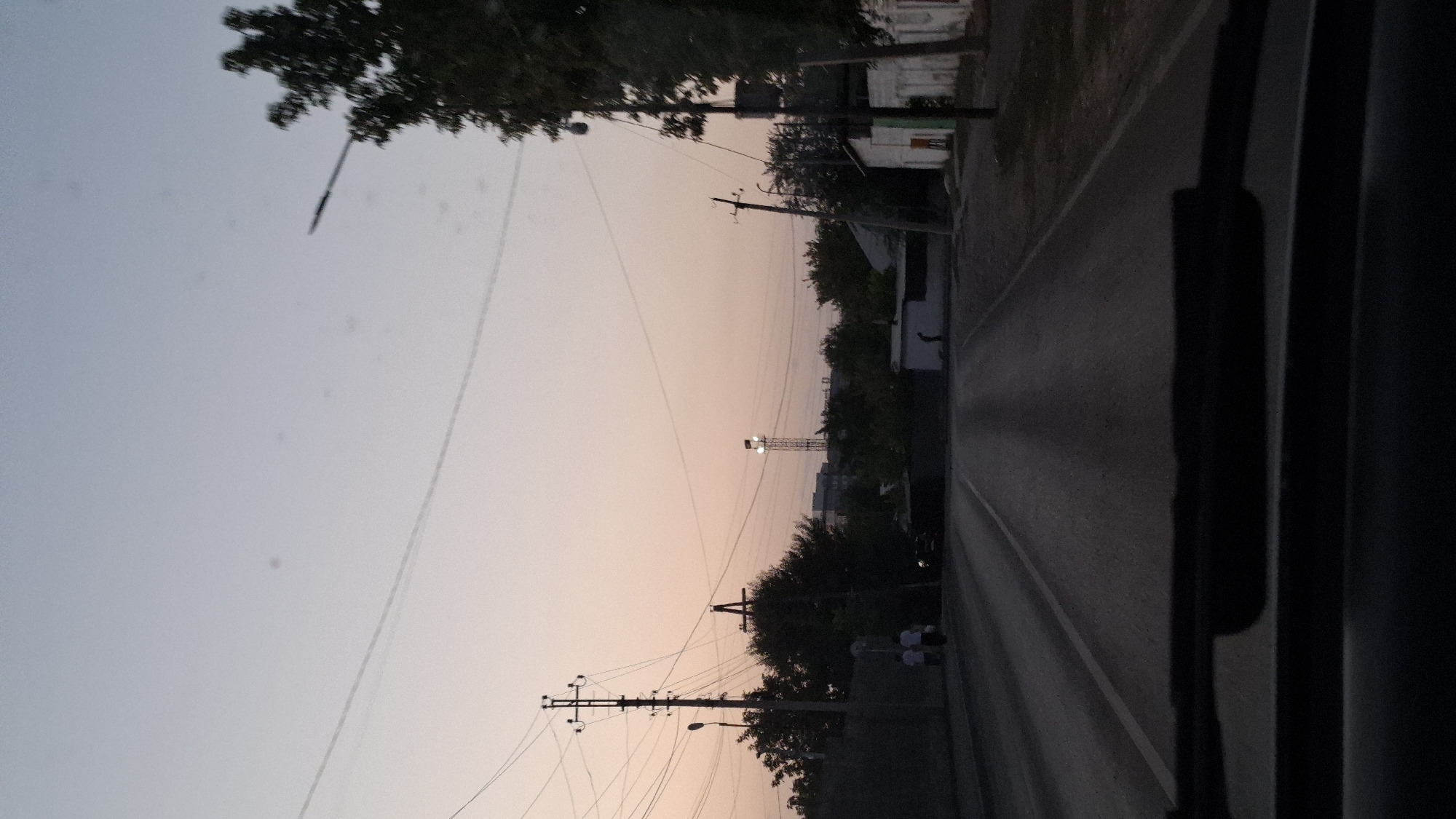 The image depicts a view from inside a vehicle driving down a road in Shymkent, Kazakhstan, at dusk. The sky is a gradient of pale blue and soft orange, indicating the setting sun. Overhead power lines crisscross the frame, supported by utility poles. On the left side of the road, there is a grassy embankment with some trees. Two individuals are walking on the embankment. Further down the road, on the right, are several buildings, including a small white structure with a green door. In the distance, a tall metal tower with a light at its apex is visible, possibly a communication tower or a street lamp. The road ahead is asphalt, with white lane markings visible. The overall impression is that of a residential or semi-industrial area on the outskirts of a city.