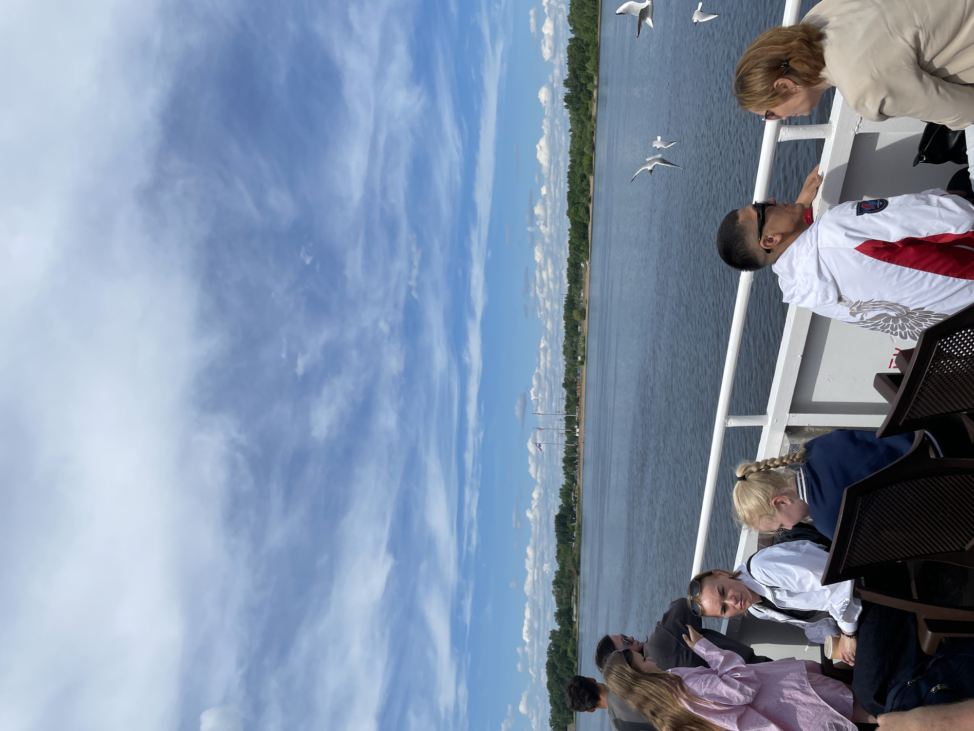 This image captures a vibrant daytime scene aboard a boat or ferry, likely on the Kama River in Perm, Russia. The perspective is from the deck, looking out towards a wide expanse of water and a distant treeline under a bright blue sky filled with scattered white clouds.
Several people are visible on the boat. In the foreground, on the right side of the frame, a man in a white and red jacket, wearing sunglasses, is leaning against the railing, looking out. Beside him, a woman in a light-colored jacket is also leaning over the railing, observing the view. Further into the boat, other passengers are seated or standing. A woman with blonde braided hair in a navy jacket is visible, as is another woman in a white jacket and sunglasses, and a woman in a pink striped shirt with long blonde hair. Parts of other individuals are also seen, suggesting a group outing.
Several white seagulls are a prominent feature, flying close to the boat and over the water, adding dynamism to the scene. The distant shoreline features a continuous treeline, with some faint structures and flags visible, indicating human activity along the riverbanks. The water appears calm, reflecting the bright sky. The weather seems pleasant and sunny, ideal for a river cruise. No discernible text is visible in the image.