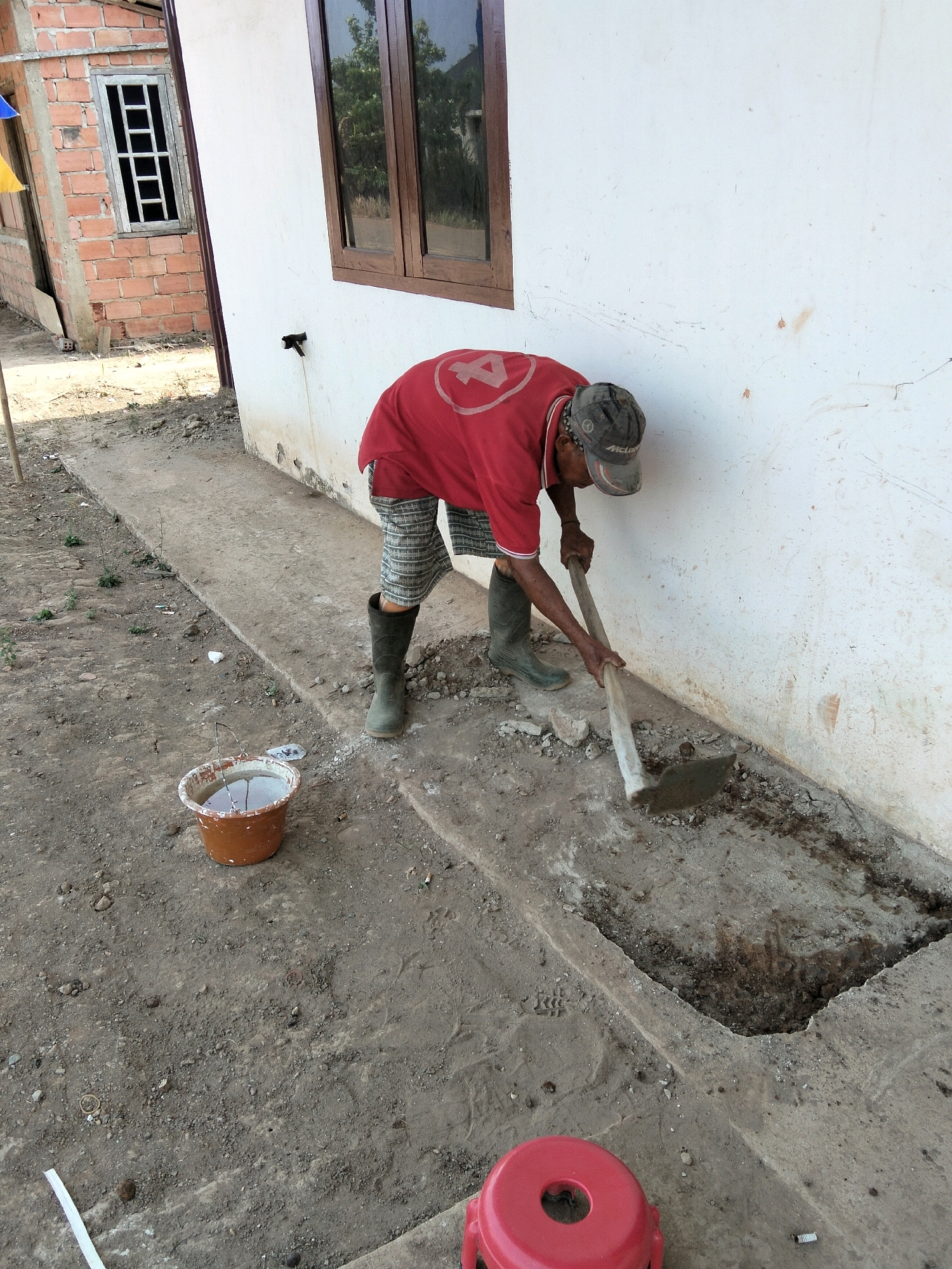 A man wearing a red t-shirt, grey and white plaid shorts, a camouflage baseball cap, and dark green rubber boots is digging a trench along the base of a white-painted building. He is using a hoe with a wooden handle and a metal blade to break up the ground. The building appears to be under construction, with exposed brickwork visible on an adjacent structure. In the foreground, to the left of the man, is a small, orange bucket filled with water. The ground is made up of dirt and small rocks. In the far foreground, a red plastic stool is visible. The scene suggests outdoor construction or repair work taking place in a residential area of Perambahan, Indonesia.