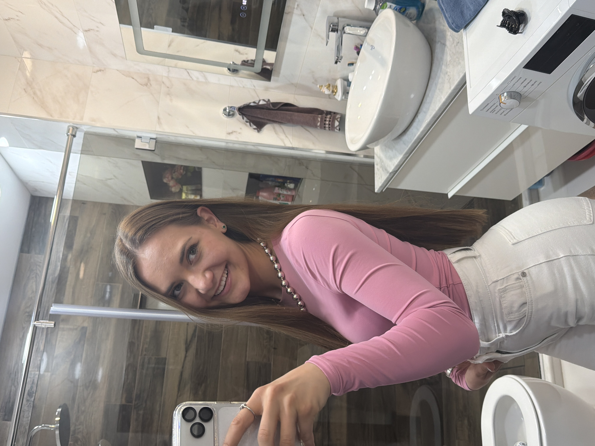 A smiling young woman captures a mirror selfie in a modern bathroom located in Buhovo, Bulgaria. She holds a smartphone, likely an iPhone given its triple-lens camera setup and clear case, in her right hand. Her long, straight brown hair frames her face as she grins widely at the camera, conveying happiness. She is dressed in a long-sleeved pink top, a pearl-like beaded necklace, and high-waisted light-colored jeans, with a ring visible on her left hand.
The bathroom features a contemporary design with elegant finishes. The wall behind her reflection is adorned with white marble-patterned tiles. A large rectangular mirror with an illuminated frame hangs above a matching marble-patterned countertop. A white circular vessel sink with a chrome faucet and various toiletries occupies the counter. A brown patterned hand towel hangs from a chrome hook on the wall. To the right of the sink area, a white washing machine with a control dial and a digital display, where some indistinct text related to settings is visible, stands next to the counter. A black hair clip is placed on top of the washing machine.
To the left of the woman's reflection, a glass shower enclosure with chrome fittings reveals dark wood-look tiles within. A white toilet is partially visible in the foreground, towards the bottom right of the image. The bathroom is brightly lit, indicative of either good natural light or effective artificial lighting, contributing to a clean and inviting atmosphere.