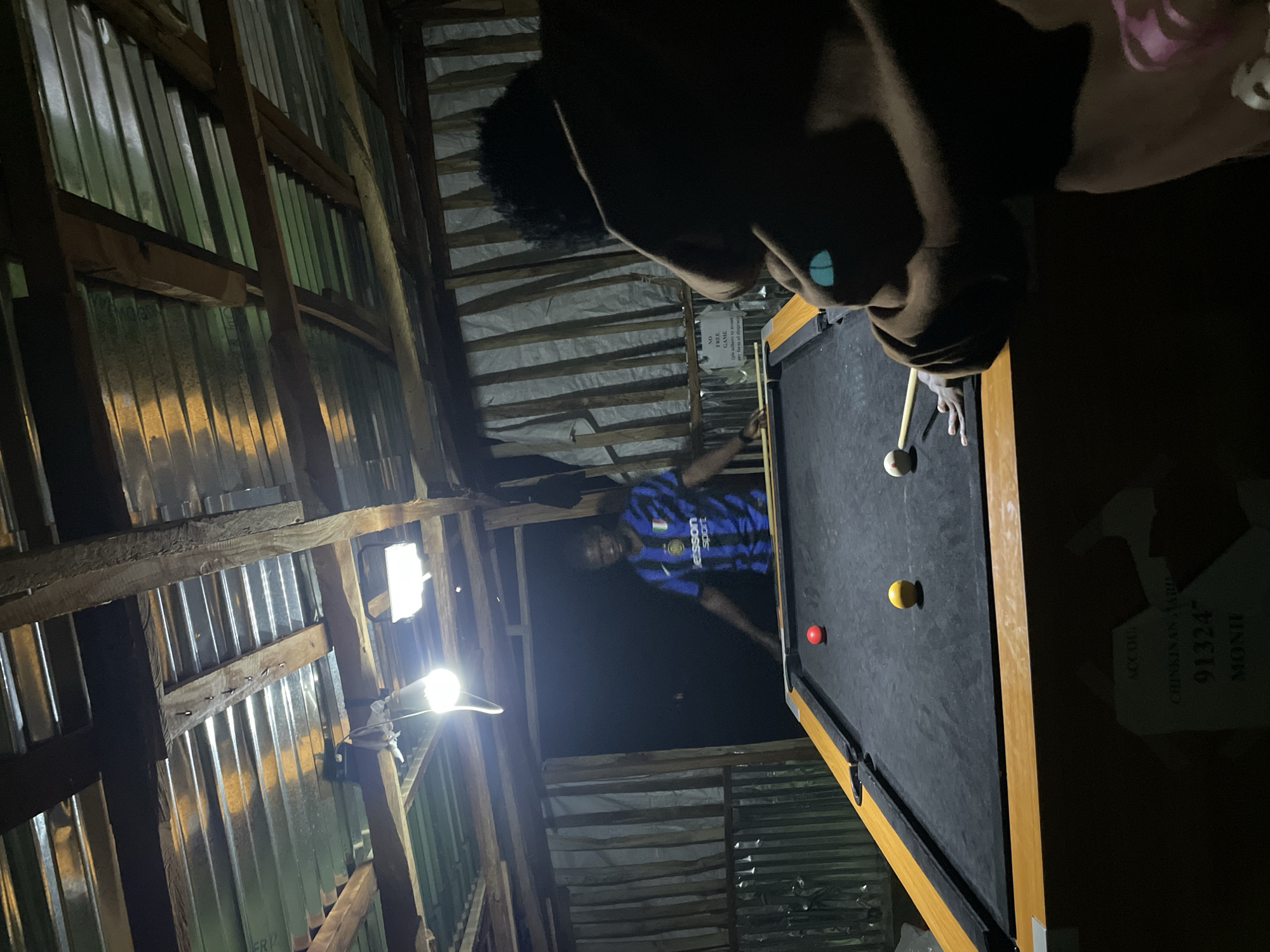 In Durumi, Nigeria, two individuals are engaged in a game of billiards. The scene takes place in a dimly lit, open-air structure with a corrugated metal roof supported by wooden beams. A pool table with a black felt surface and wooden frame is centrally located. On the table, several billiard balls are visible: a red ball, a yellow ball, and a white cue ball. One individual, wearing a blue and black striped soccer jersey, stands behind the pool table, facing it. The other individual, partially obscured and facing away from the camera, is leaning over the table, seemingly about to take a shot. The background consists of a corrugated metal wall and wooden planks. A sign, possibly a notice or advertisement, is attached to the wall, though its text is difficult to discern. The overall ambiance is casual and recreational.