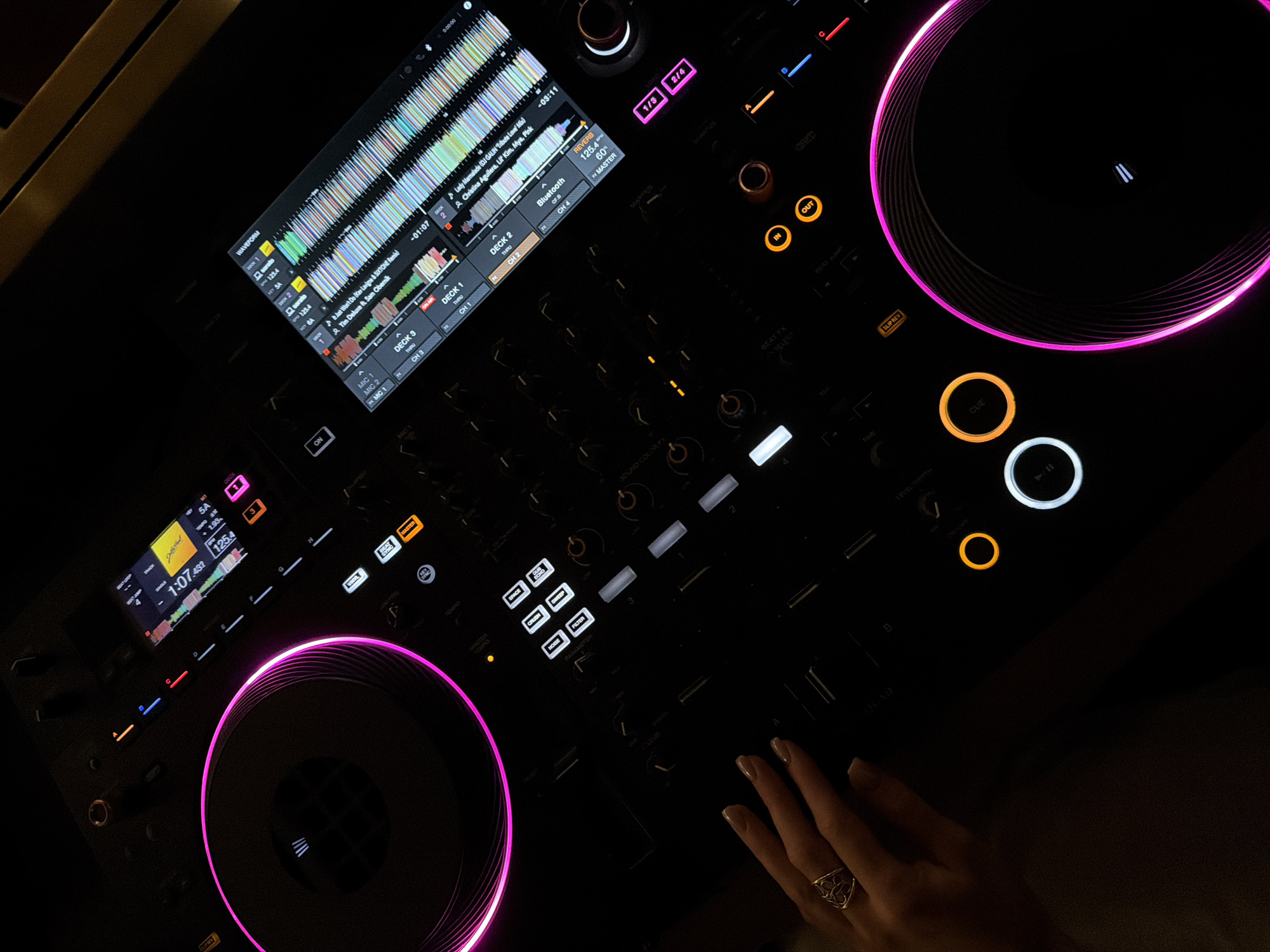 In Budva, Montenegro, a close-up, high-angle shot captures a DJ's setup in a dimly lit environment. The focus is on a black DJ controller with illuminated elements. A woman's left hand, adorned with a silver ring and nude-colored fingernails, rests on the lower left portion of the controller, fingers extended over some of the buttons. The DJ controller features two prominent circular jog wheels, one on the left and one on the right, both outlined with vibrant pink LED lighting.
The central area of the controller displays a touchscreen interface showing waveform visualizations for multiple audio decks (DECK 1, DECK 2, DECK 3, and DECK 4). Song titles like "Hot Won't Do (Leipzig & ISSTOMR remix)" and "Lady Marmalade (DJ Gain & Tiësto Remix)" are visible on the screen. Various buttons and knobs are present across the controller, some illuminated in different colors, including orange, blue, and white. A few buttons are labeled with functions such as "ON," "REVERSE," "PHASER," "FLANGER," "KEY," and "CUE."
The background is dark, with hints of muted wooden textures visible at the top right, suggesting a possible bar or club interior. The overall ambiance is professional and focused on the DJ equipment.