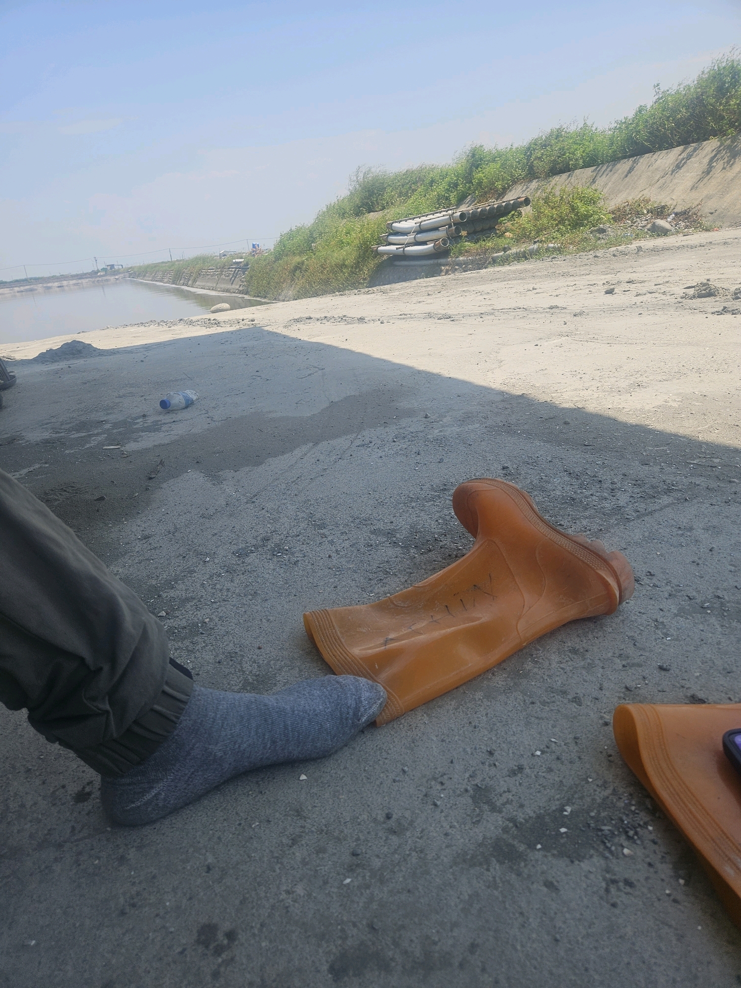 An individual wearing a gray sock and dark green pants sits on a dirt path next to a body of water. An orange rubber boot lies on the ground in front of them. In the background, a concrete embankment is covered in green vegetation, with a pile of pipes stacked on top. A plastic water bottle is visible on the ground to the left. The scene is illuminated by daylight under a clear sky. The location is Taixi Township, Taiwan.