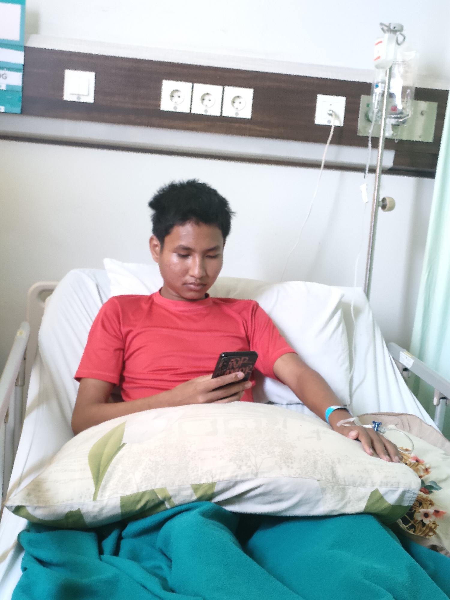 A young man with dark hair is lying in a hospital bed, looking at his phone. He is wearing a red t-shirt and a blue wristband. An IV drip is connected to his arm. A pillow with a green leaf pattern is resting on his lap, and a teal blanket is draped over his legs. The hospital room has white walls and a wooden panel with electrical outlets above the bed. An IV pole is visible to the right of the bed. The scene appears to be in Pekanbaru, Indonesia, as indicated by context clues that are not visually present in the image itself.