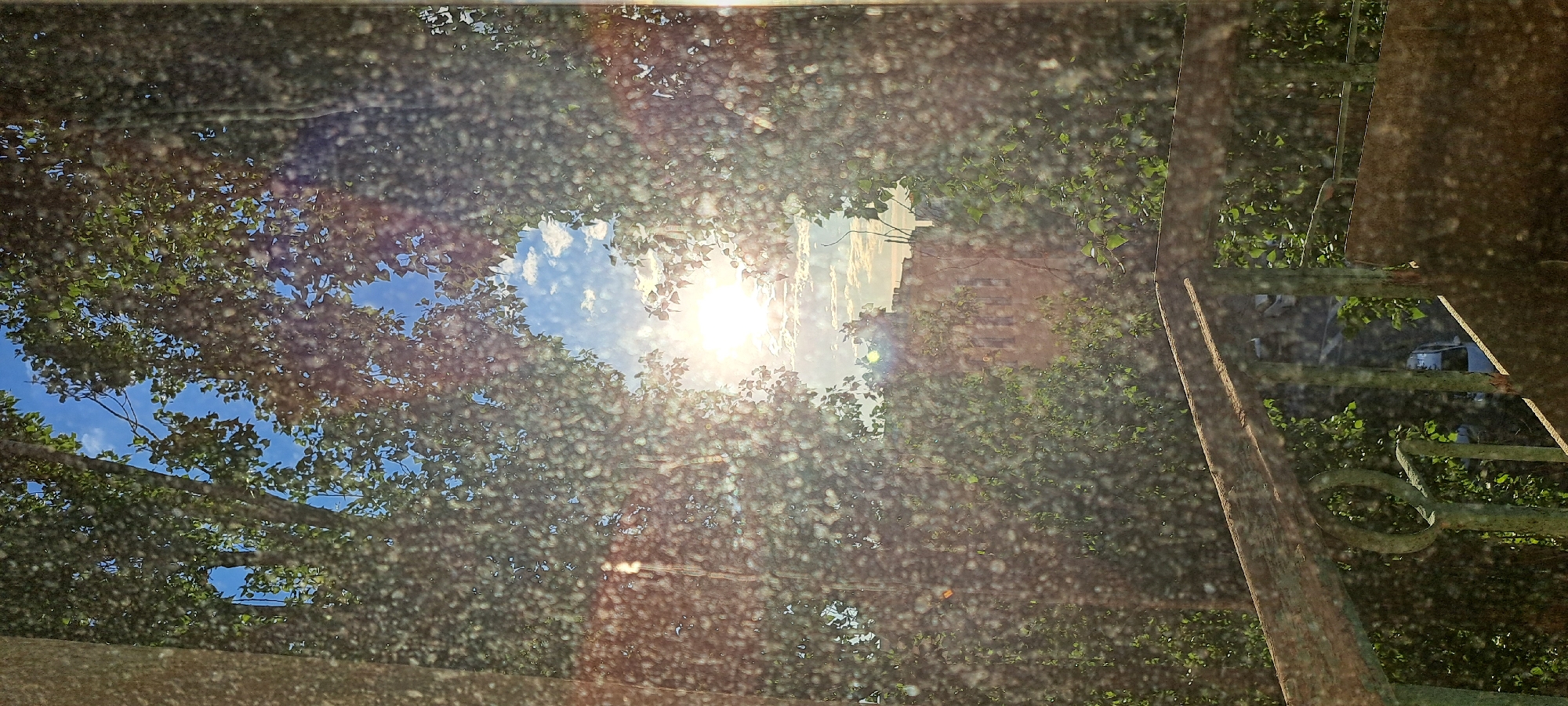 The image depicts a view through a window or glass surface covered in many small water droplets or smudges, obscuring parts of the scene. The foreground features a weathered, rusty metal railing, possibly part of a balcony or staircase, with green paint peeling. Beyond the railing, lush green foliage from trees fills the sides of the frame, creating a natural frame. In the center, a bright sun is visible in a blue sky dotted with white clouds. A building with a reddish-brown facade and visible windows is partially seen through the foliage in the background. The overall impression is an outdoor scene, possibly urban, viewed from a protected vantage point.