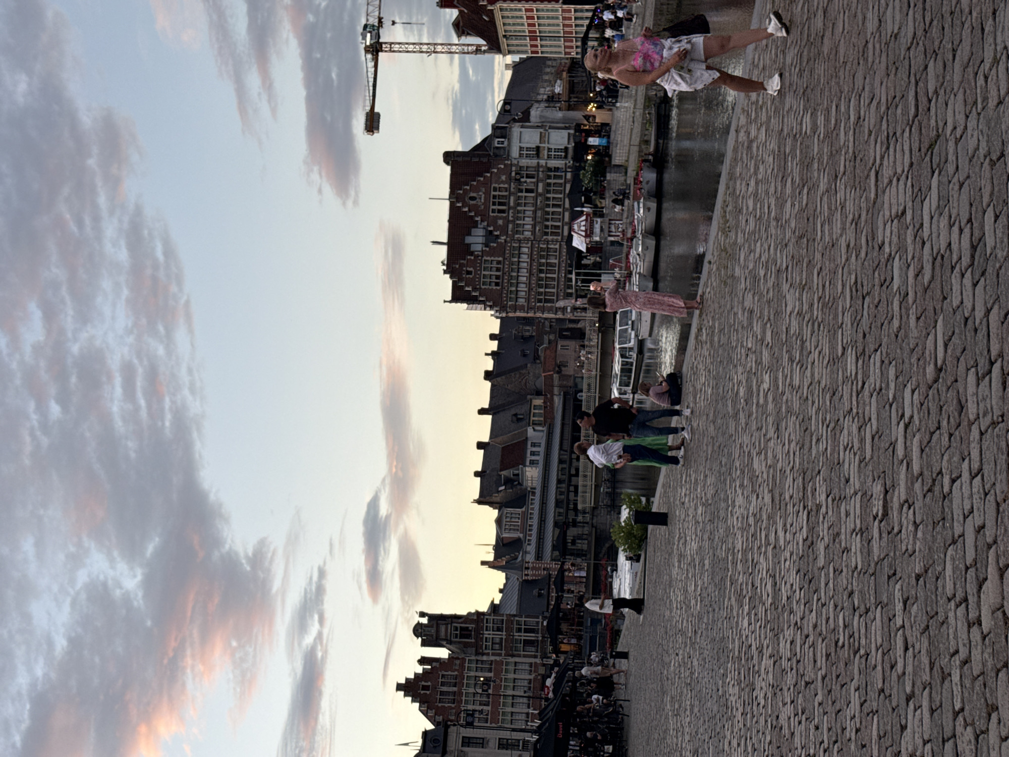 The image depicts a canal-side promenade in Ghent, Belgium, likely the Graslei or Korenlei. The sky is filled with soft, pastel-colored clouds, suggesting either dawn or dusk. A cobblestone path occupies the foreground, with several people walking and standing along the embankment.
On the left side, several people are seated at tables outside what appears to be a cafe or restaurant, with awnings providing shade. A few individuals are standing near the water's edge. In the mid-ground, a group of four men are standing on the promenade. To their right, a woman in a pink dress stands by the canal, appearing to take a photo with her phone. Further to the right, a woman in white shorts and a floral top walks across the cobblestones, carrying a dark bag.
The canal itself is lined with boats, and on the opposite bank, historical buildings with distinctive gabled roofs characteristic of Ghent architecture are visible. A construction crane is prominent in the upper right portion of the image, indicating ongoing development or renovation in the area. The overall scene conveys a relaxed, urban atmosphere with a blend of historical and modern elements.