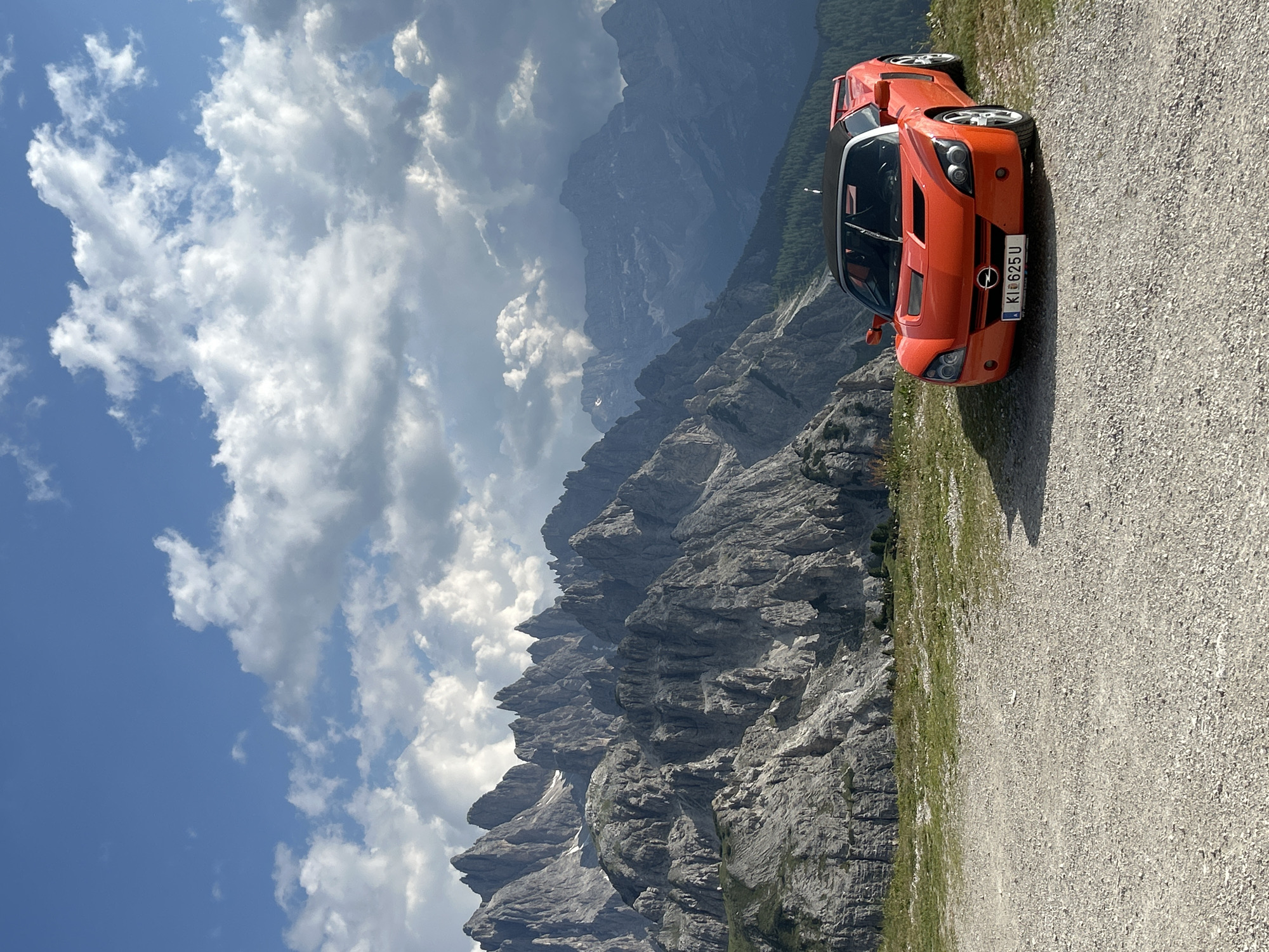 This is a landscape photograph taken from an elevated viewpoint overlooking a dramatic mountain range. In the foreground, parked on a gravelly path, is a bright orange sports car, possibly an Opel Speedster or Vauxhall VX220, with its license plate reading "KI 625 U". The car is angled towards the viewer, showcasing its sleek design.
The background is dominated by rugged, grey mountain peaks, which appear weathered and jagged, suggesting a high-altitude alpine environment. The scale of the mountains is immense, with layers of ridges fading into the distance. A strip of green grass borders the gravel path next to the car, hinting at a summer or late spring setting.
The sky is a vibrant blue, dotted with scattered, fluffy white clouds, indicating fair weather. The lighting suggests it is daytime, likely midday or early afternoon, with the sun casting some shadows but generally illuminating the scene. The overall mood is one of adventure and freedom, with the car positioned as if ready to embark on a scenic drive through the mountains. The image is oriented vertically.