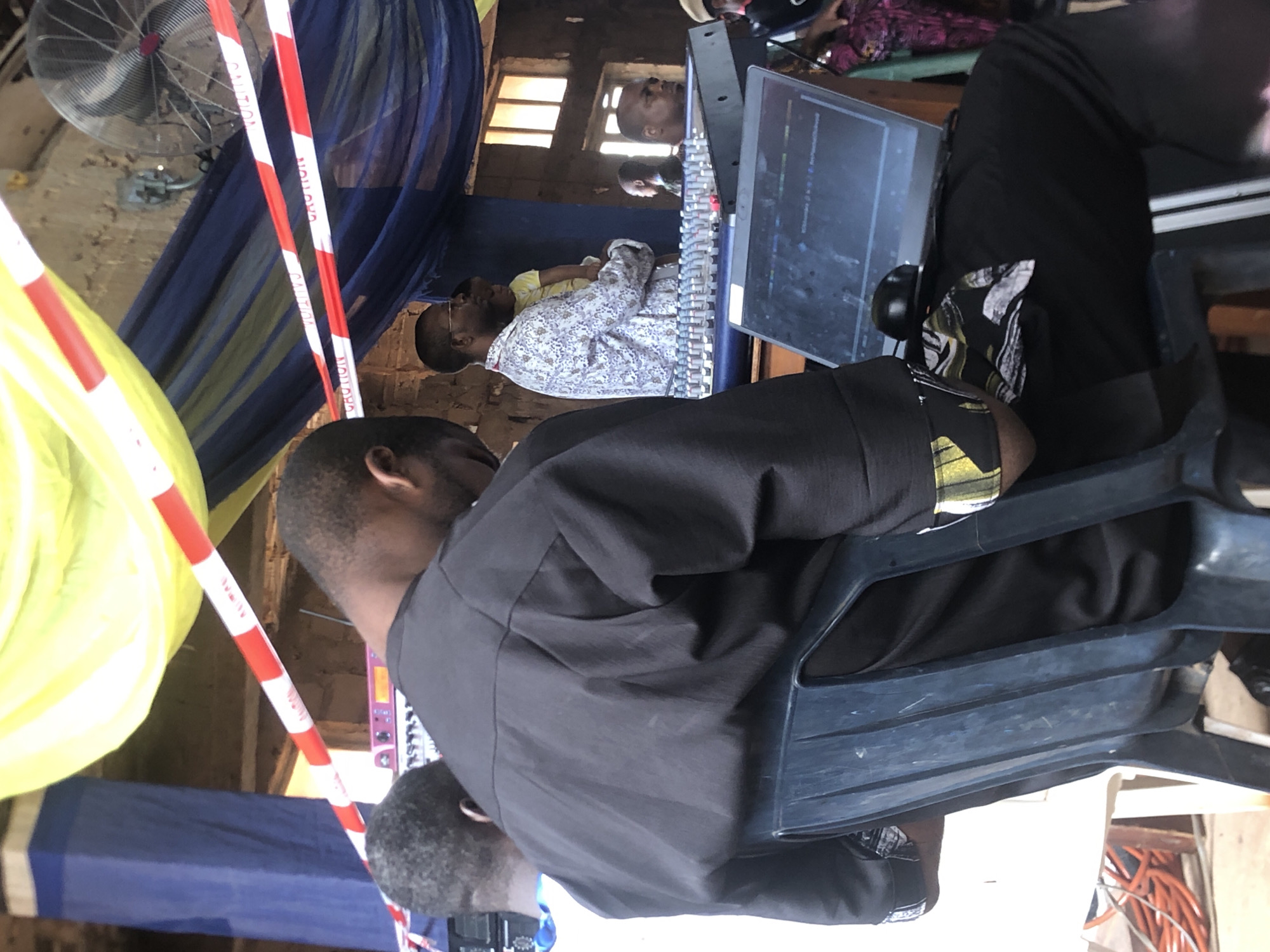 The image captures an indoor scene at what appears to be a small event venue in Obosi, Nigeria. A man wearing a black shirt is seated on a blue plastic chair, facing a mixing console with a computer screen. Behind him, another man in a floral shirt sits at a similar console, and a third individual can be seen in a window reflection. Yellow and blue decorative cloths hang from the ceiling, along with caution tape. A fan is visible near the ceiling. The location appears somewhat cluttered.