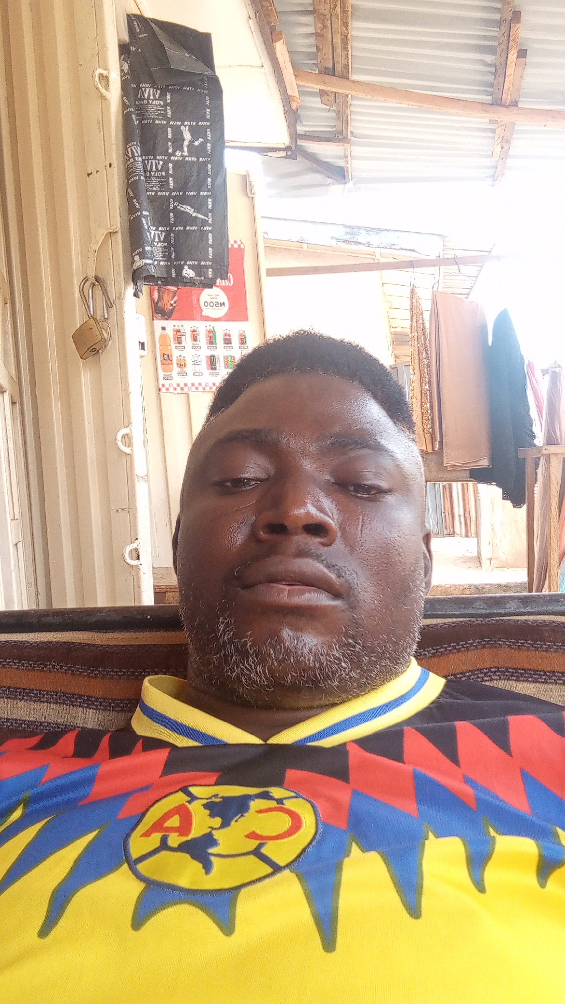 A close-up selfie shows a man wearing a yellow Club America soccer jersey with blue and red details. The background includes a corrugated metal structure with an advertisement, a padlock hanging from a handle, and clothing hanging on a rack. The man appears to be in the city of Sabon Tasha, Nigeria, based on the branding on the advertisement.