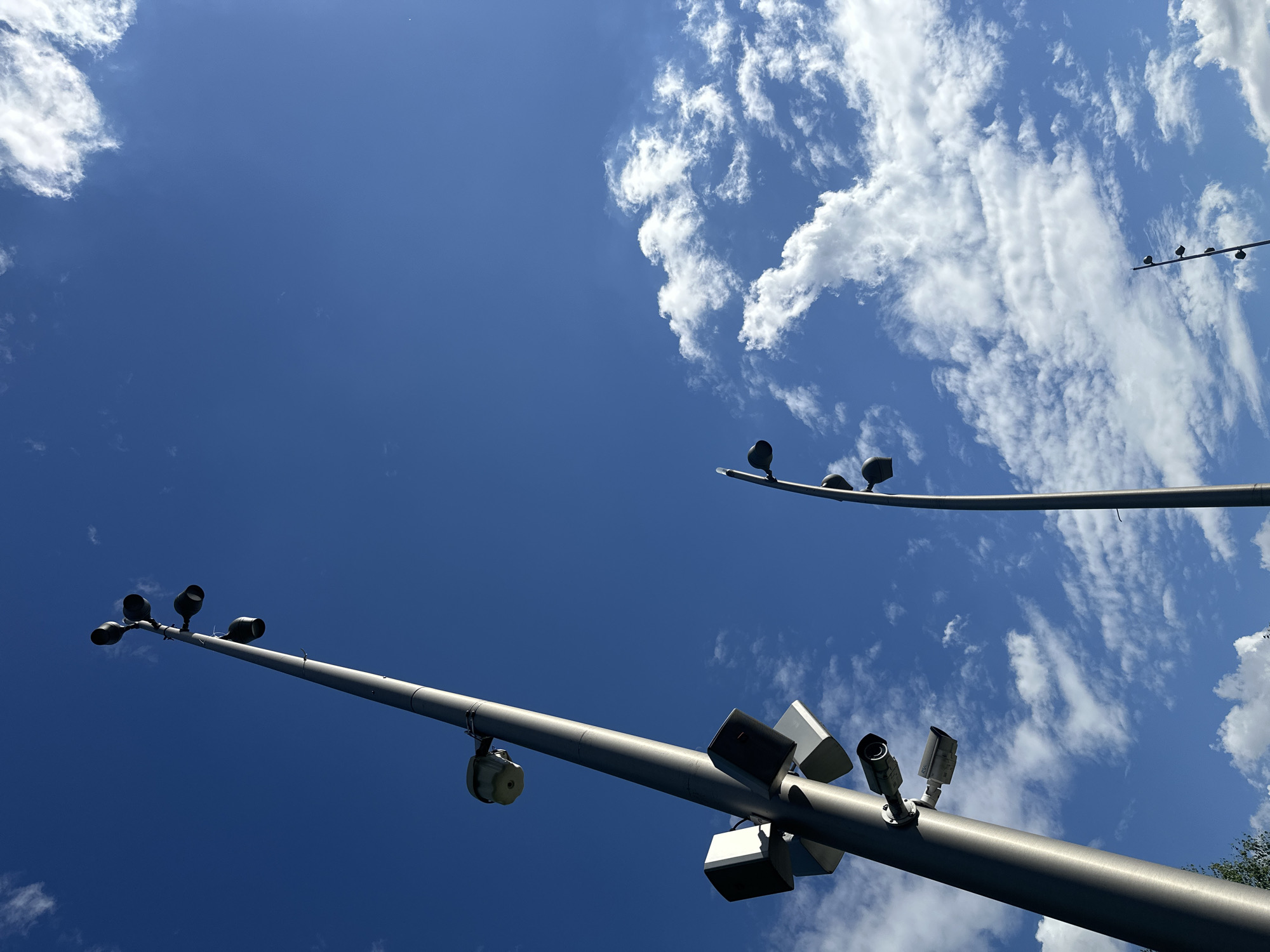 The media file shows a low-angle view of a clear blue sky with scattered white clouds. Several poles are visible, extending upwards and angled across the frame. The poles are equipped with various devices, including what appear to be surveillance cameras and possibly lights or speakers. One pole in the foreground has multiple cameras and box-like objects attached. Another pole further in the background also has several camera-like devices. The scene suggests an outdoor public space, possibly a street or park, equipped with monitoring or lighting systems. The lighting and cloud formations suggest it is daytime, likely mid-morning or afternoon. No people are visible in the frame. There is no visible text in the image. The overall impression is one of security or public infrastructure against a backdrop of a bright, partly cloudy sky. Given the location context of Moscow, Russia, this could be a scene from a public area in the city.
