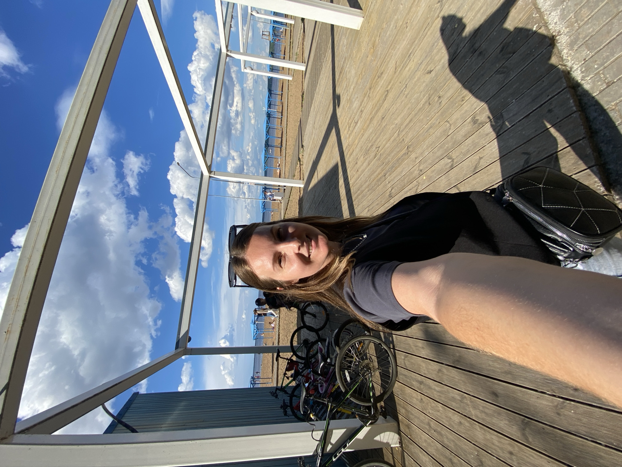 This media file depicts a woman taking a selfie outdoors on a sunny day. She is seated on a wooden deck, with a beach and body of water visible in the background. Several bicycles are parked behind her, suggesting a recreational area or a place where people cycle. The sky is blue with fluffy white clouds, indicating fair weather. The sun is casting strong shadows, suggesting it is either morning or late afternoon. The scene appears to be a beach or a promenade in Chaikovsky, Russia, given the context. The woman is smiling, conveying a sense of happiness or contentment. She is wearing a dark t-shirt and sunglasses. A black handbag is placed next to her. There is no visible text in the media file.