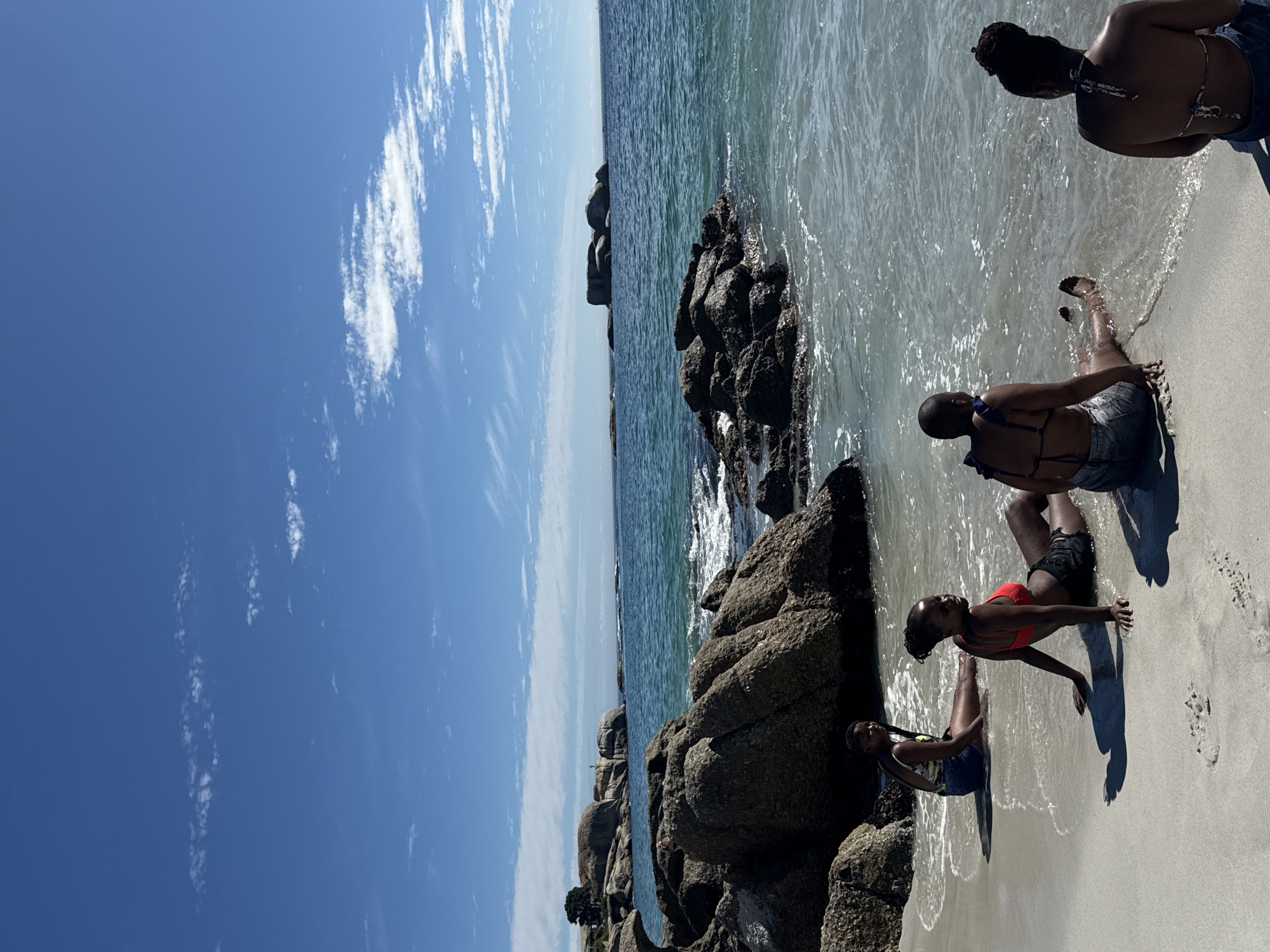 The image displays Boulders Beach in Simon's Town, Cape Town, South Africa, on a clear, sunny day. The sky is blue with scattered white cumulus and cirrus clouds. Four adult females are seated on the light-colored, wet sand at the ocean's edge. From right to left: one female is positioned with her back to the viewer, wearing a patterned halter-neck bikini top and dark shorts; immediately to her left, another female also faces away from the viewer, wearing a dark blue bikini top and light denim shorts, with her feet immersed in the water. Further left, a third female is seated facing left, wearing a red bikini top and dark shorts. The fourth female, positioned further left, is seated facing slightly away from the camera, wearing a patterned tank top and denim shorts. Gentle waves lap at the individuals' legs and the shoreline. The environment is characterized by numerous large, rounded granite boulders distributed along the beach and extending into the clear, turquoise ocean. In the distant background to the left, additional large boulders, some green vegetation, and a single light-colored building are visible on the coastline, along with a small, indistinct human figure standing on a distant rock outcrop.