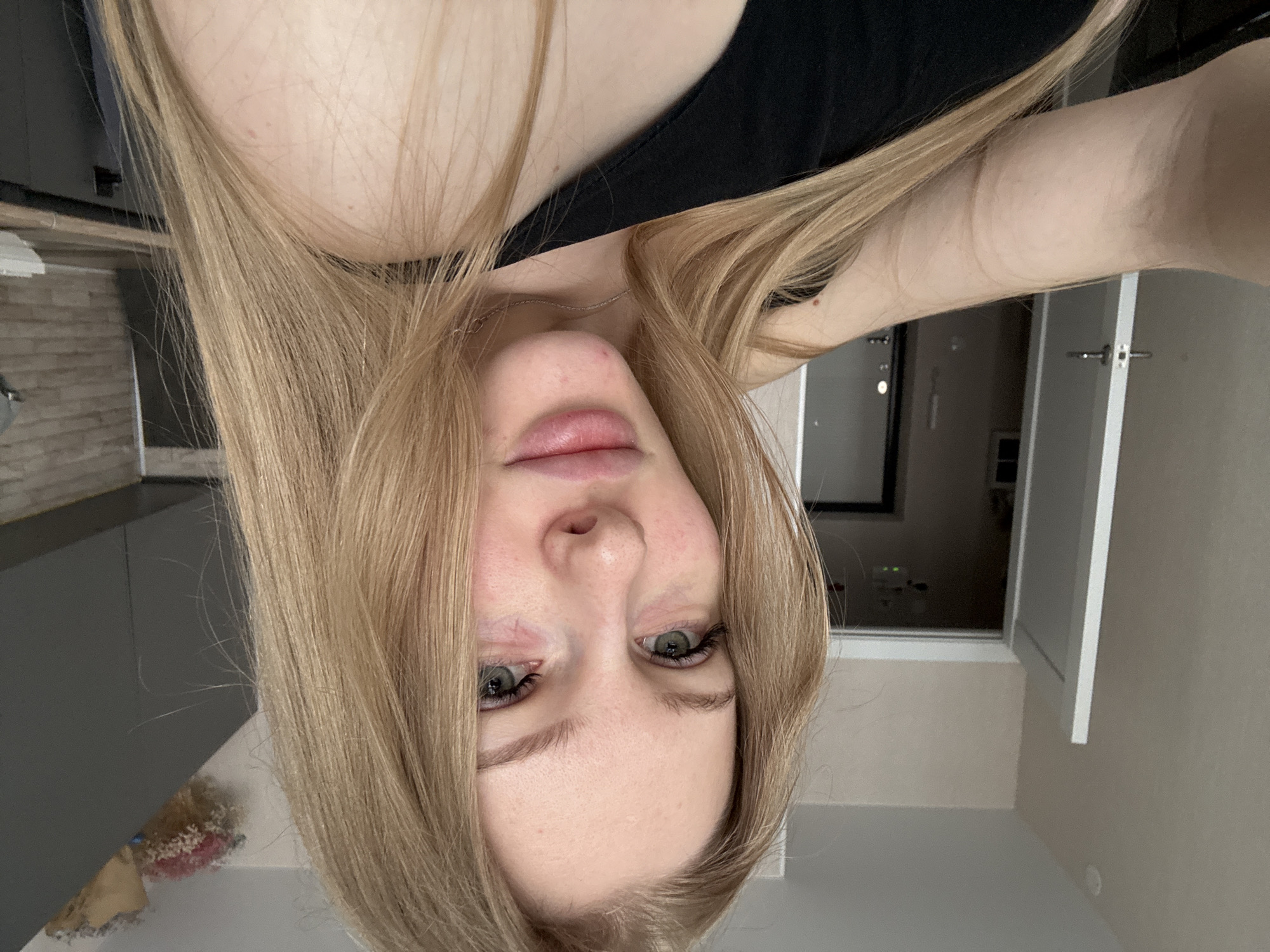 The image shows a blonde woman with fair skin, green eyes, and a black tank top. The photo is taken from a close, upside-down angle, focusing on her face. The background includes a glimpse of a kitchen with gray cabinets, a bouquet of flowers on a countertop, and a door. Based on image analysis and available data, there is no information to suggest that the image was taken in the city of Perm, Russia. Therefore, a concise, forensic, matter-of-fact description of the scene does not include the location in the city of Perm, Russia.