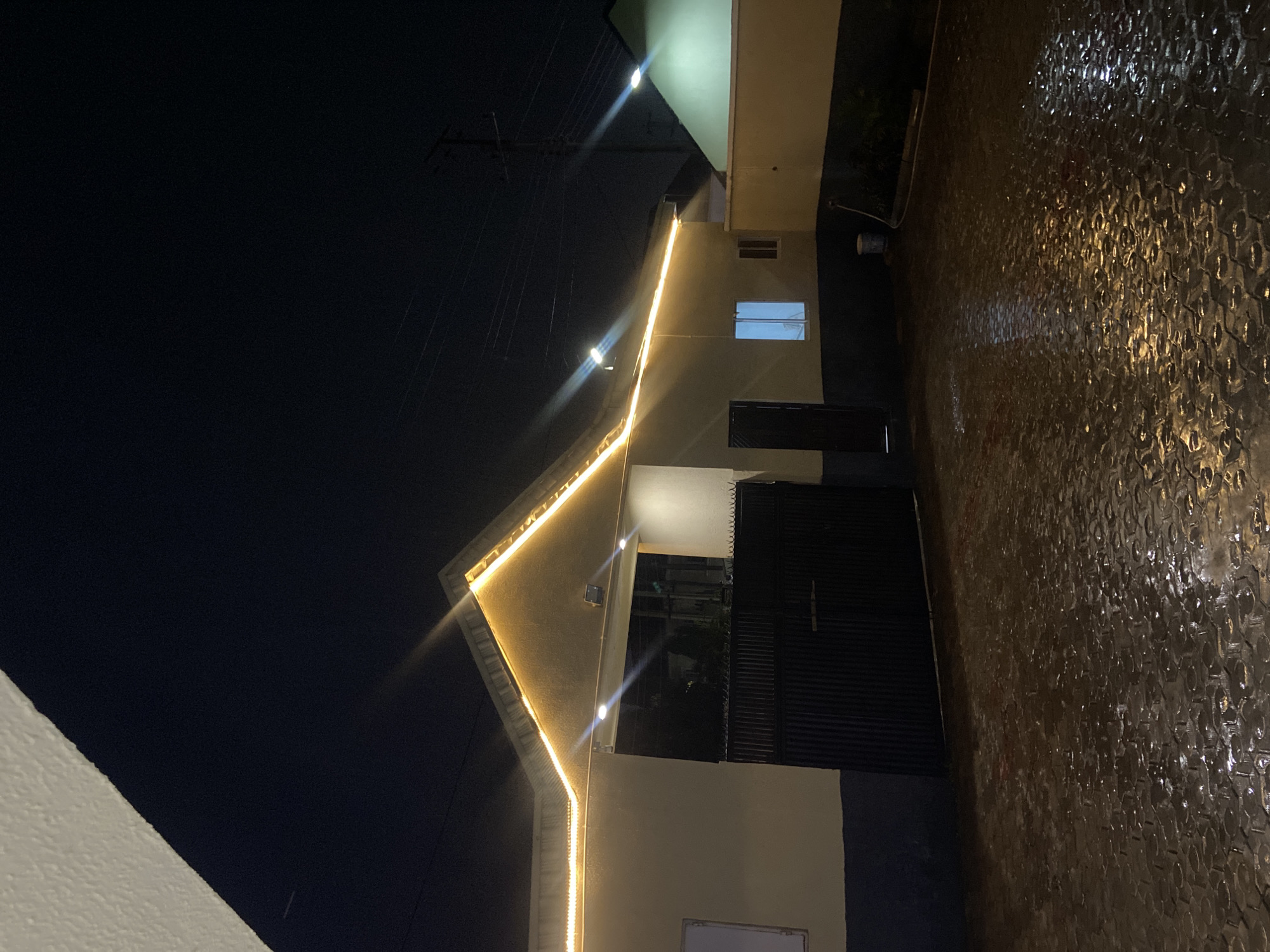 The image depicts a night scene in Gura Top, Nigeria, featuring the exterior of a building illuminated by festive lights along the roofline and exterior lighting. The ground in front of the building is paved with interlocking stones and appears wet, reflecting the surrounding lights, suggesting recent rain or a damp environment. A dark, corrugated metal gate is visible, and to the right, there's a wall and another section of the building with a lit window. The sky is dark, indicating nighttime. There are no people visible in the scene.