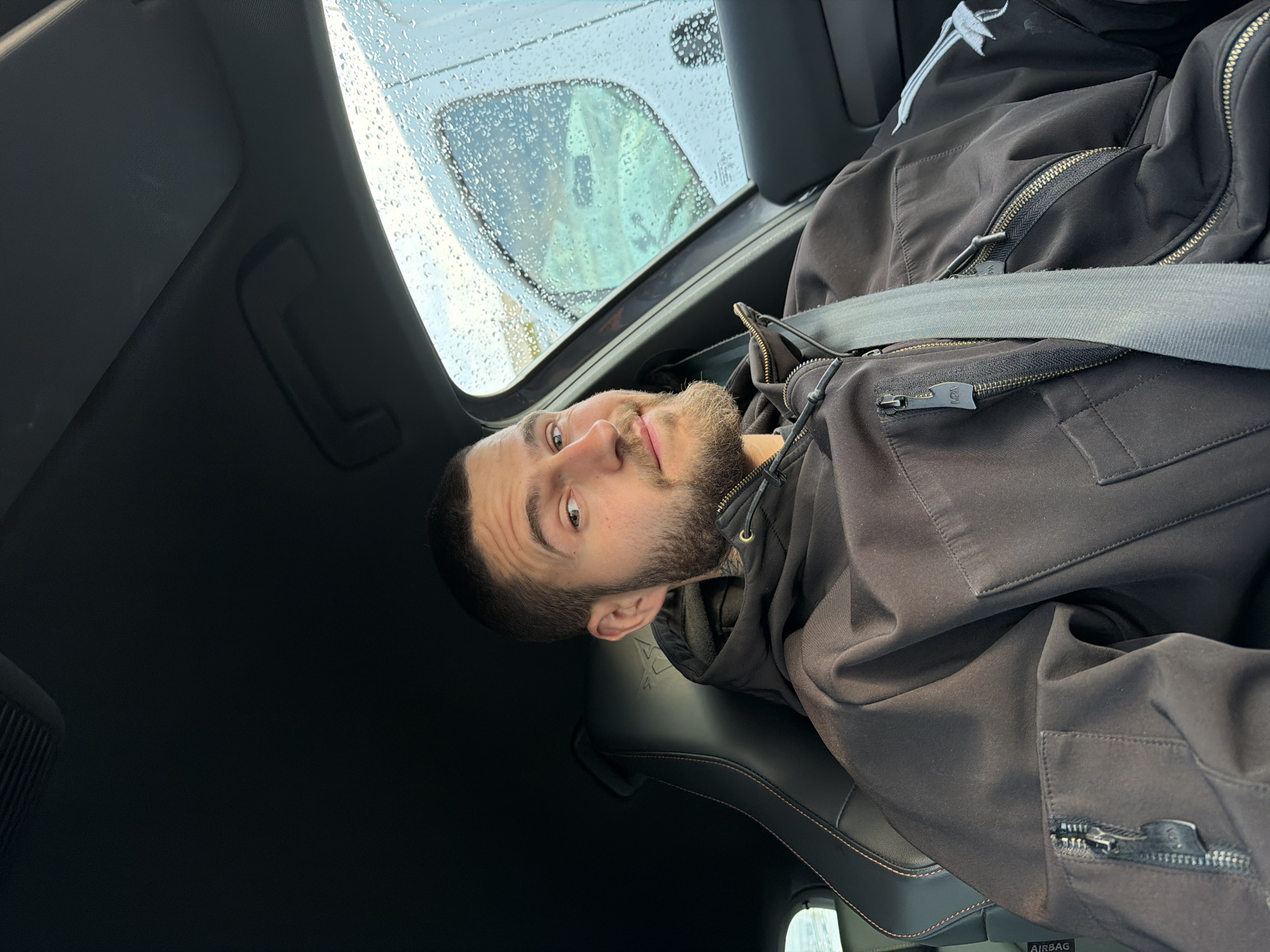 A bearded man is seated in a car, wearing a dark jacket and a seatbelt. Raindrops are visible on the sunroof. The car interior is black with visible seat stitching. The location is unknown and cannot be determined from the image.