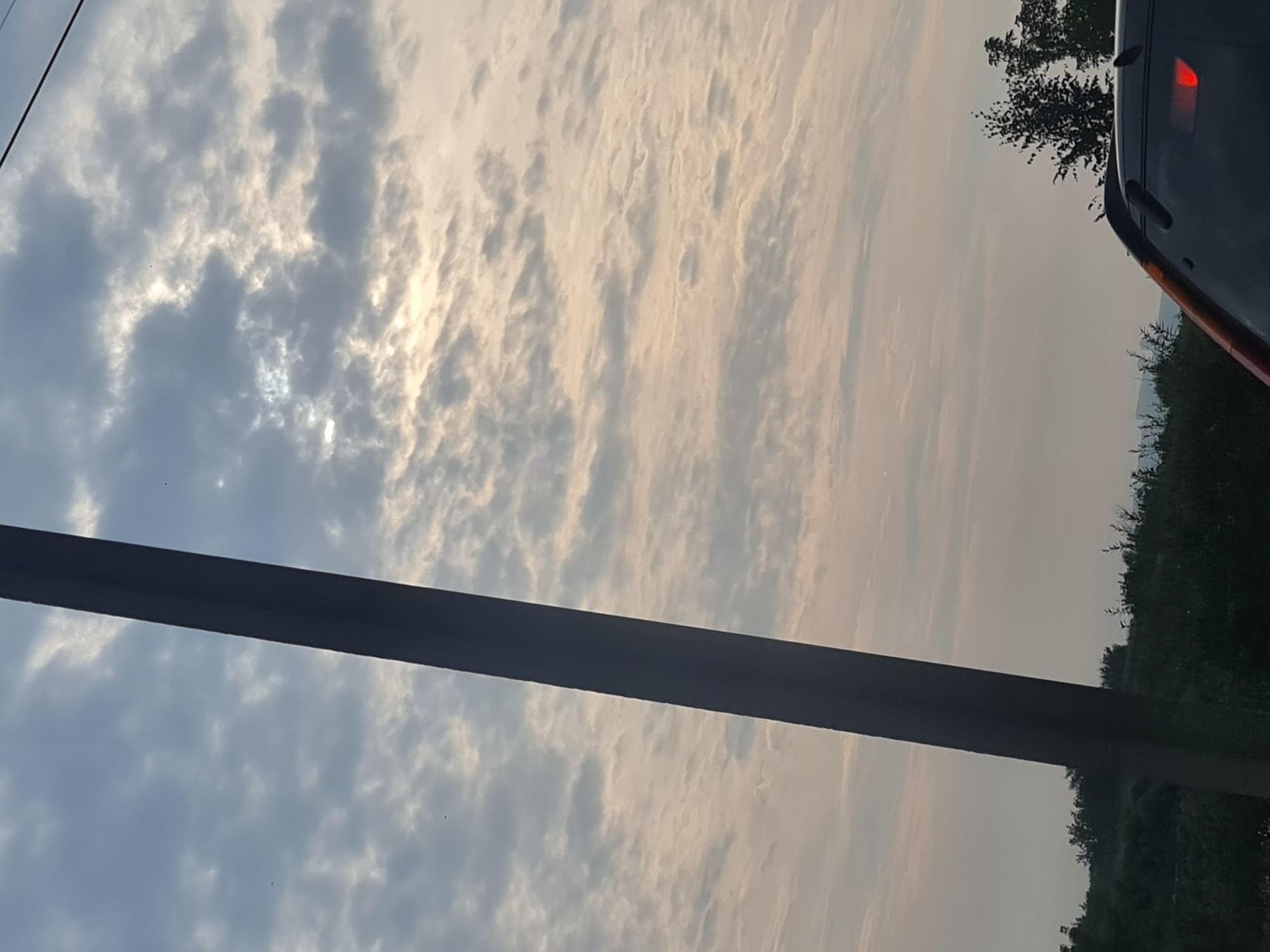 The image depicts a cloudy sky with the sun partially visible through the clouds. A dark pole is positioned on the left side of the frame, running vertically. In the lower portion of the image, there is a glimpse of a dark-colored car with its red brake light illuminated, suggesting it might be stationary or in traffic. A line of dark green trees and vegetation forms a horizon line beneath the sky. The overall scene appears to be an outdoor environment, possibly on the outskirts of a city or in a rural area, with a muted color palette due to the cloudy conditions. There is no information in the image that would allow for identification of the city of Irkutsk, Russia.