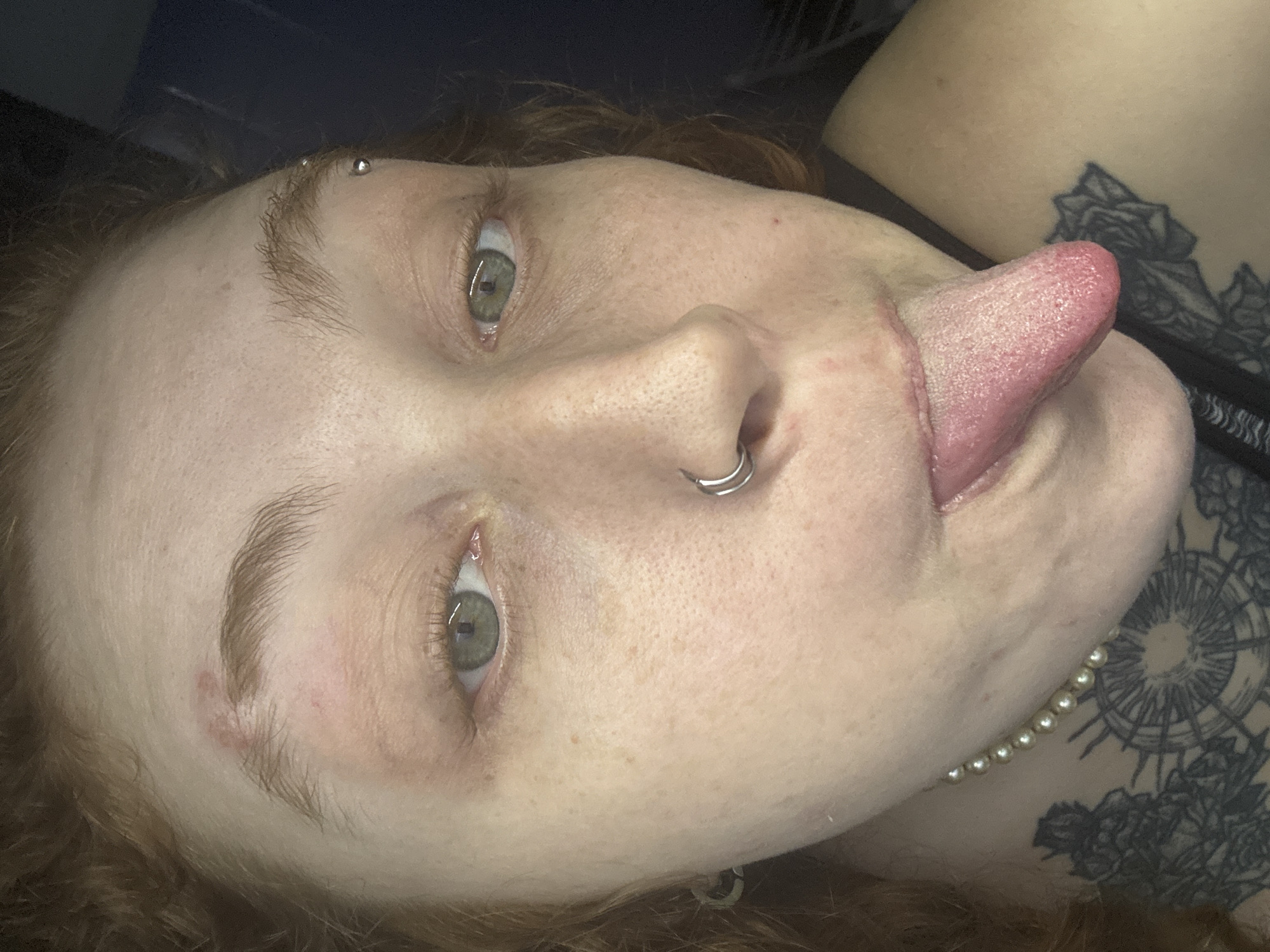 A close-up portrait shows a person lying down, looking towards the camera, with their tongue playfully extended to the right side of their face. The person has fair skin with visible freckles and reddish-brown, curly hair that frames their face. Their eyes are a striking greenish-grey.
Visible body modifications include a silver hoop septum piercing and an eyebrow piercing with two silver balls above their left eye. A reddish patch is present on the skin beneath the left eyebrow. Around their neck, a pearl necklace is partially visible. On the upper chest and shoulder area, a black ink tattoo featuring intricate, possibly floral or mandala-like patterns, is prominent.
The background is dark and indistinct, suggesting an indoor setting. The person's expression appears casual and relaxed. This scene is set in Buenos Aires, Argentina.