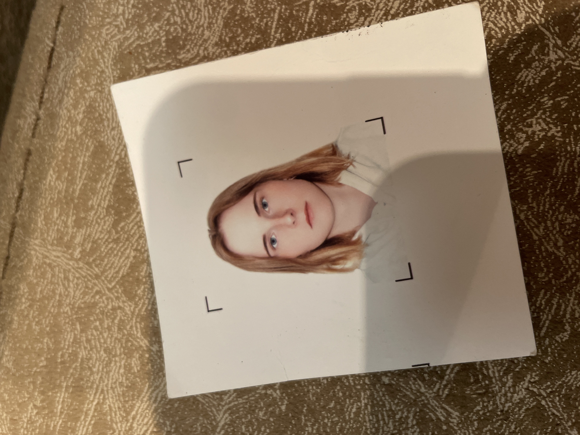 A passport-style photograph of a young woman with shoulder-length blonde hair and blue eyes is centered on a white card. She is wearing a light-colored hooded garment. The card has small, black corner registration marks. The card is placed on a textured, beige fabric surface. The photograph appears to be taken indoors.