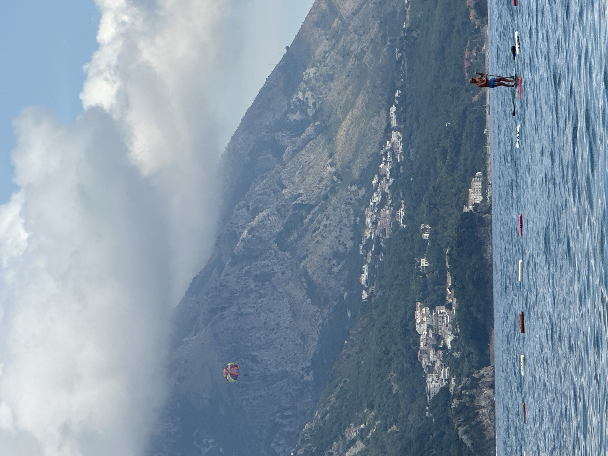An outdoor, eye-level, full shot captures a man on a paddleboard in the Adriatic Sea off the coast of Boreti, Montenegro. The man, wearing blue shorts, stands on a paddleboard, holding a paddle. A series of buoys stretch across the foreground of the water. In the background, a mountainous landscape is visible, dotted with white buildings. A colorful parasail floats in the sky above the mountains. The sky is partly cloudy, with patches of blue peeking through.