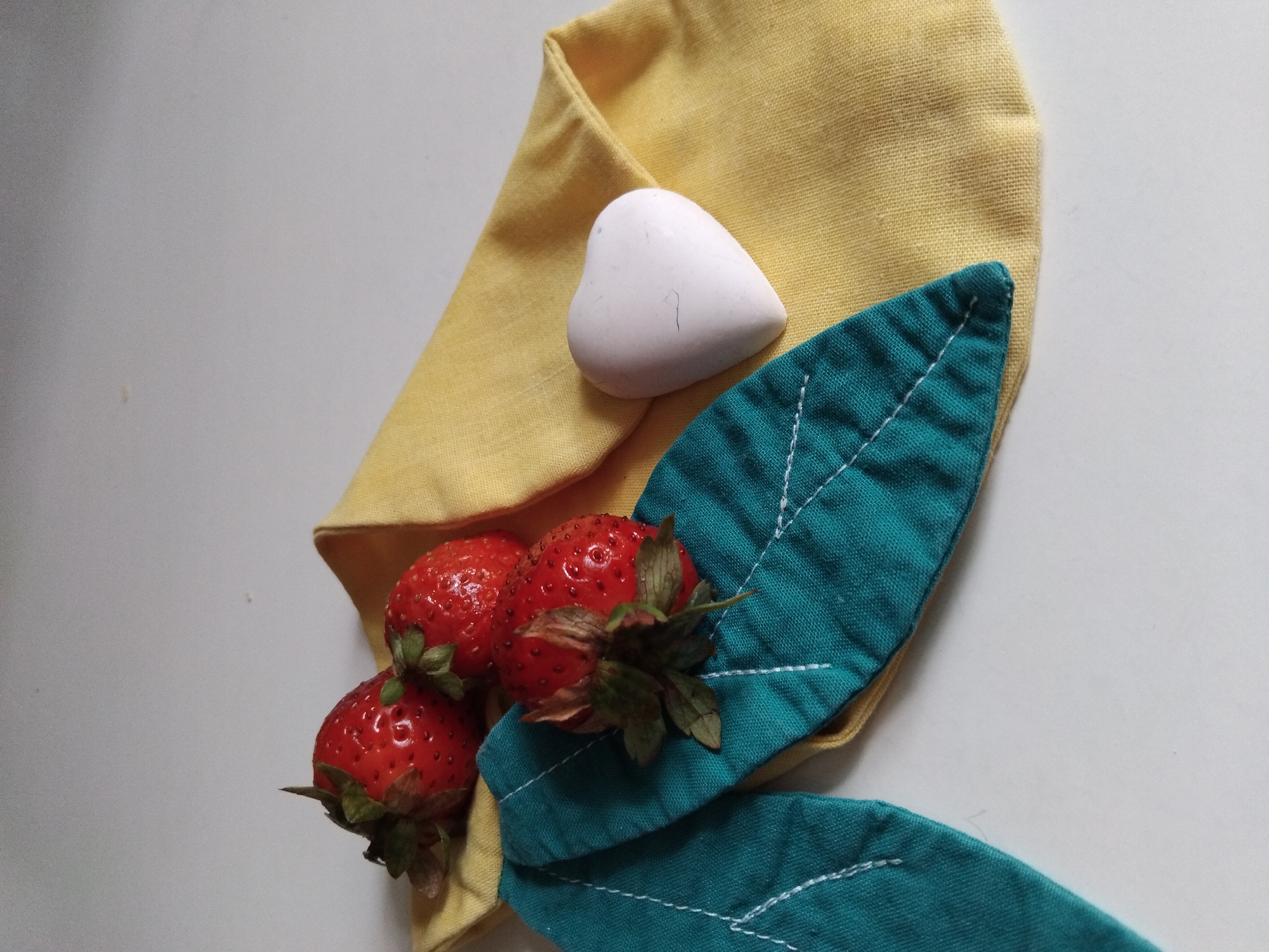 The image shows a still life arrangement on a white surface. Three ripe strawberries are clustered together, resting on a folded yellow fabric. To the left of the strawberries, two teal fabric cutouts shaped like leaves with white stitching are placed. A small, white heart-shaped object is positioned on the yellow fabric, to the right of the strawberries. The background is a plain, light gray surface. There is no indication of the image being located in Vila Nova de Famalicão, Portugal.