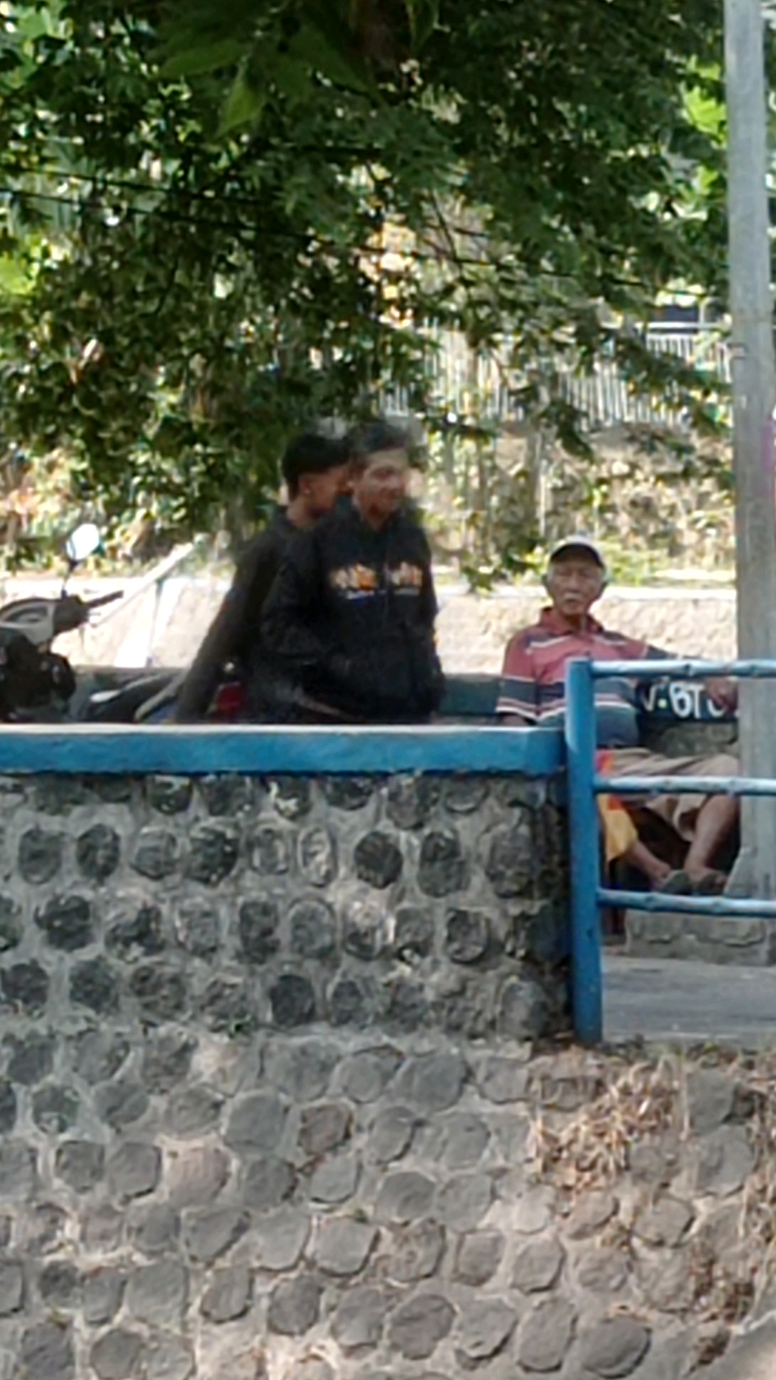 The image shows a scene outdoors, with three men present. Two men are standing behind a low stone wall, and one is sitting on the other side of the wall. The two standing men are partially obscured by trees and foliage, but they appear to be young adults. The man on the right is wearing a black hooded sweatshirt with an orange graphic on it. The man on the left is wearing dark clothing. The man sitting on the other side of the wall is an elderly man wearing a striped polo shirt and a baseball cap. He is seated on a bench or low structure behind the wall, and a portion of a blue railing is visible in front of him. A motorcycle is parked to the left of the standing men, partially visible. The wall in the foreground is constructed of irregularly shaped stones. The background shows vegetation and some distant structures that are out of focus. The scene appears to be a public area, possibly a park or a roadside rest stop in Mulyoagung, Indonesia.
