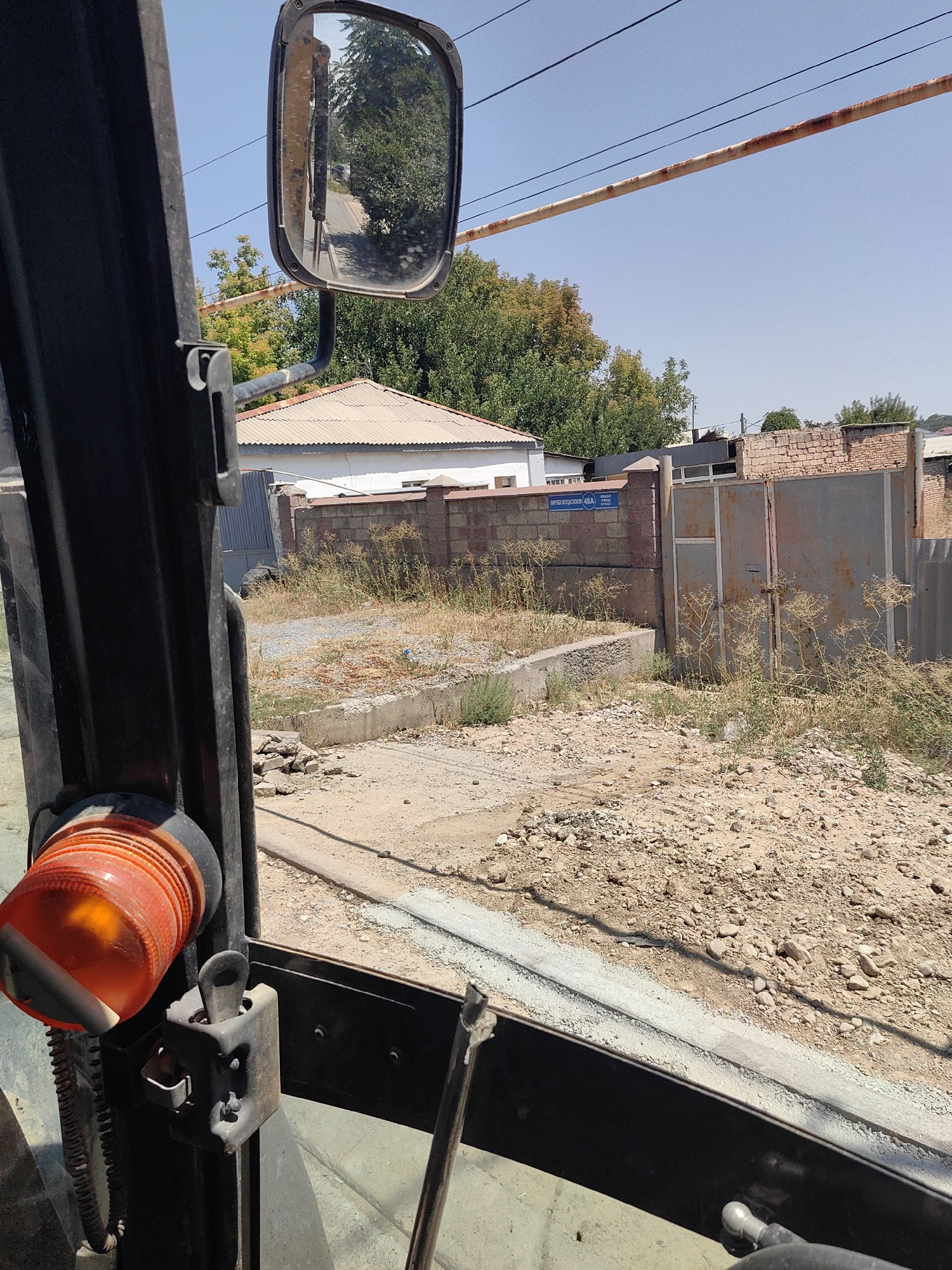 The image provides a view from inside a vehicle, looking out towards a street scene in Shymkent, Kazakhstan. A prominent feature is the vehicle's side mirror, reflecting a road and trees. In the background, there are buildings with corrugated metal roofs and brick walls, suggesting a residential or industrial area. A large metal gate is visible, and dry vegetation is scattered across the dusty ground. A street sign with text in Cyrillic and numbers is affixed to a brick wall. The overall environment appears to be dry and sunny.