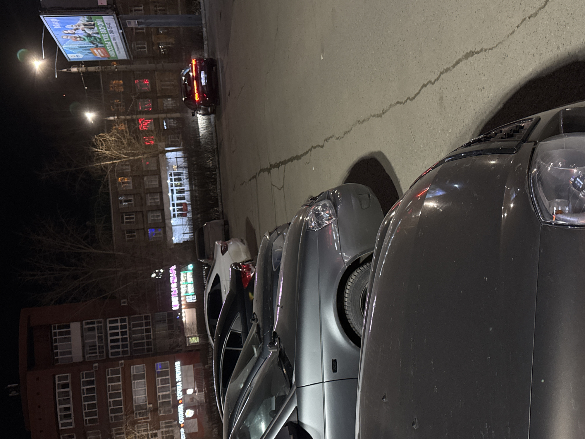The image depicts a street scene in Perm, Russia, at night. A line of parked cars stretches into the distance along the right side of the frame. A red car is visible further down the street. Buildings line the left side of the street, some with illuminated signs written in Russian. One sign reads "Цветы & Шары." Another sign displays a billboard. Streetlights provide illumination. The ground is asphalt.