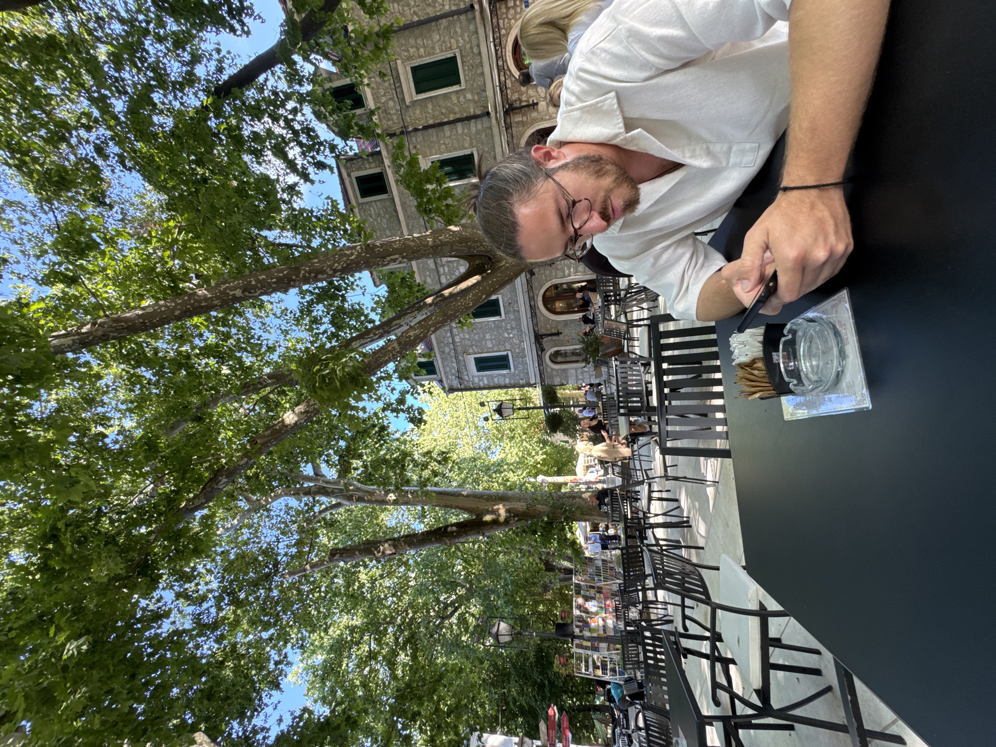 In Trebinje, Bosnia and Herzegovina, a man with glasses and a man bun, wearing a white shirt, sits at a black table in an outdoor cafe. He is looking at his phone. The table has an ashtray and a holder with toothpicks. Behind him, there are other tables and chairs, a stone building with green shutters, and large trees with green leaves. The sky is visible through the branches of the trees. In the background, there are other people sitting at tables and walking around.
