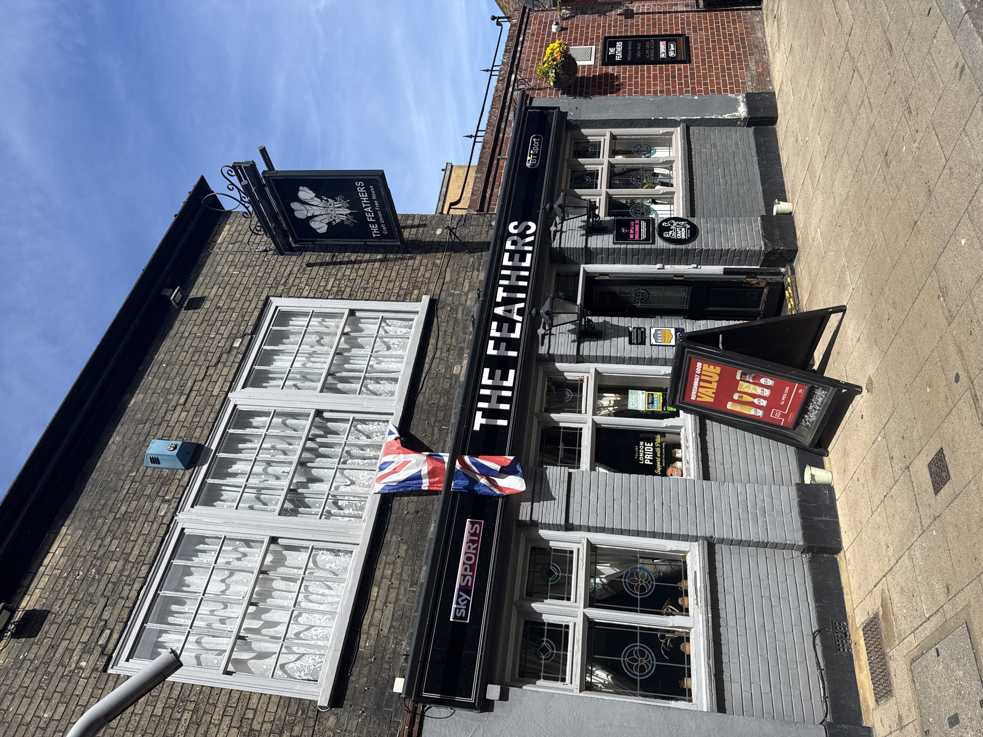 The image shows the exterior of "The Feathers," a pub located in Great Yarmouth, United Kingdom. The pub is a two-story building with a gray brick facade and white-framed windows, behind which are lace curtains. A large black sign with white lettering above the entrance displays the name "THE FEATHERS" and logos for Sky Sports and BT Sport, indicating sports coverage. A Union Jack flag is draped above the sign. A smaller, hanging sign shows the pub's name and describes it as a "Craft Union Free House." Near the entrance, an A-frame sign advertises "REFRESHINGLY GOOD VALUE" with images of beer and prices. There are signs in the windows for "FULLER'S LONDON PRIDE" and "WE OPERATE CHALLENGE 25." The pub is on a narrow street with a sidewalk. A brick building is visible behind the pub. The sky is blue and clear.