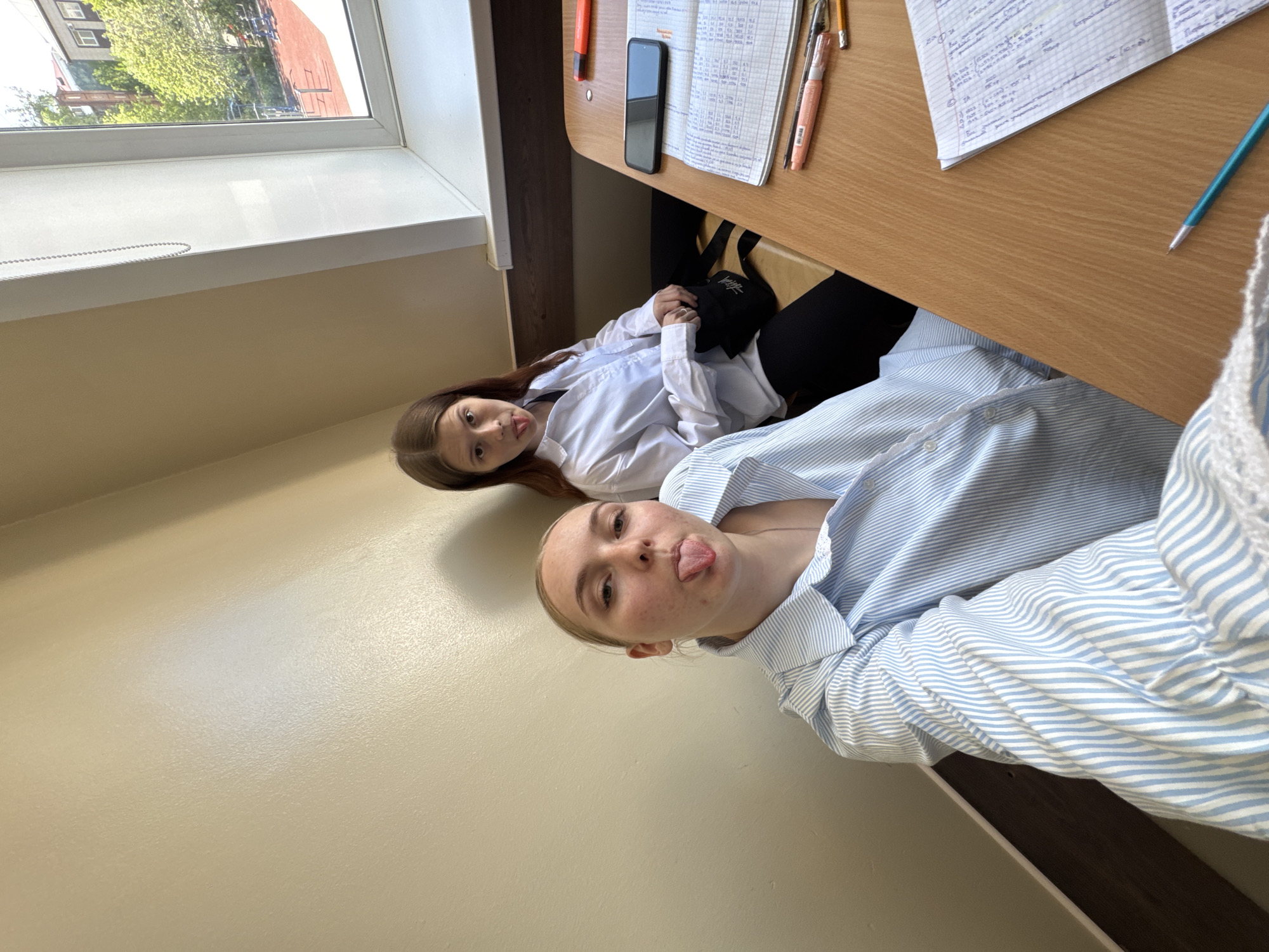 Two females are seated at a desk in what appears to be a classroom setting. One female is sticking her tongue out at the camera and is wearing a blue and white striped shirt. The other female is looking at the camera. Her blouse is white. There is a window with a view of a building and trees outside. The desk has a mobile phone, writing tools, and papers. The location is Perm, Russia.