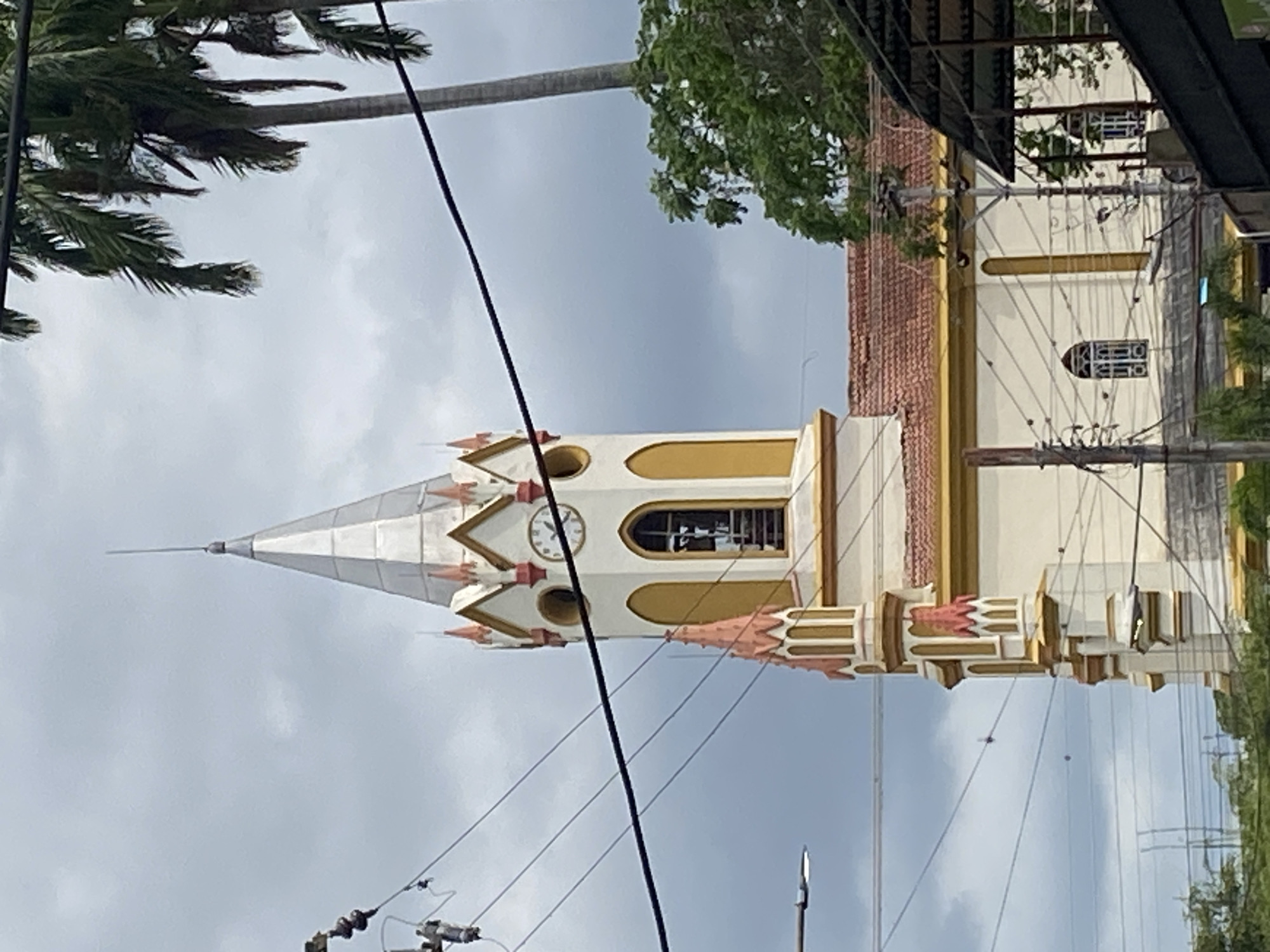 A partial view of Iglesia Inmaculada Concepción, a Catholic church in Tinaquillo, Venezuela, is visible against an overcast sky. The church's steeple, featuring a silver conical roof and a clock face, is prominent. The facade of the church is cream with yellow accents and terracotta-colored decorative elements. Utility poles and power lines run across the foreground, partially obscuring the view. Vegetation, including palm trees and other foliage, are visible in the periphery.