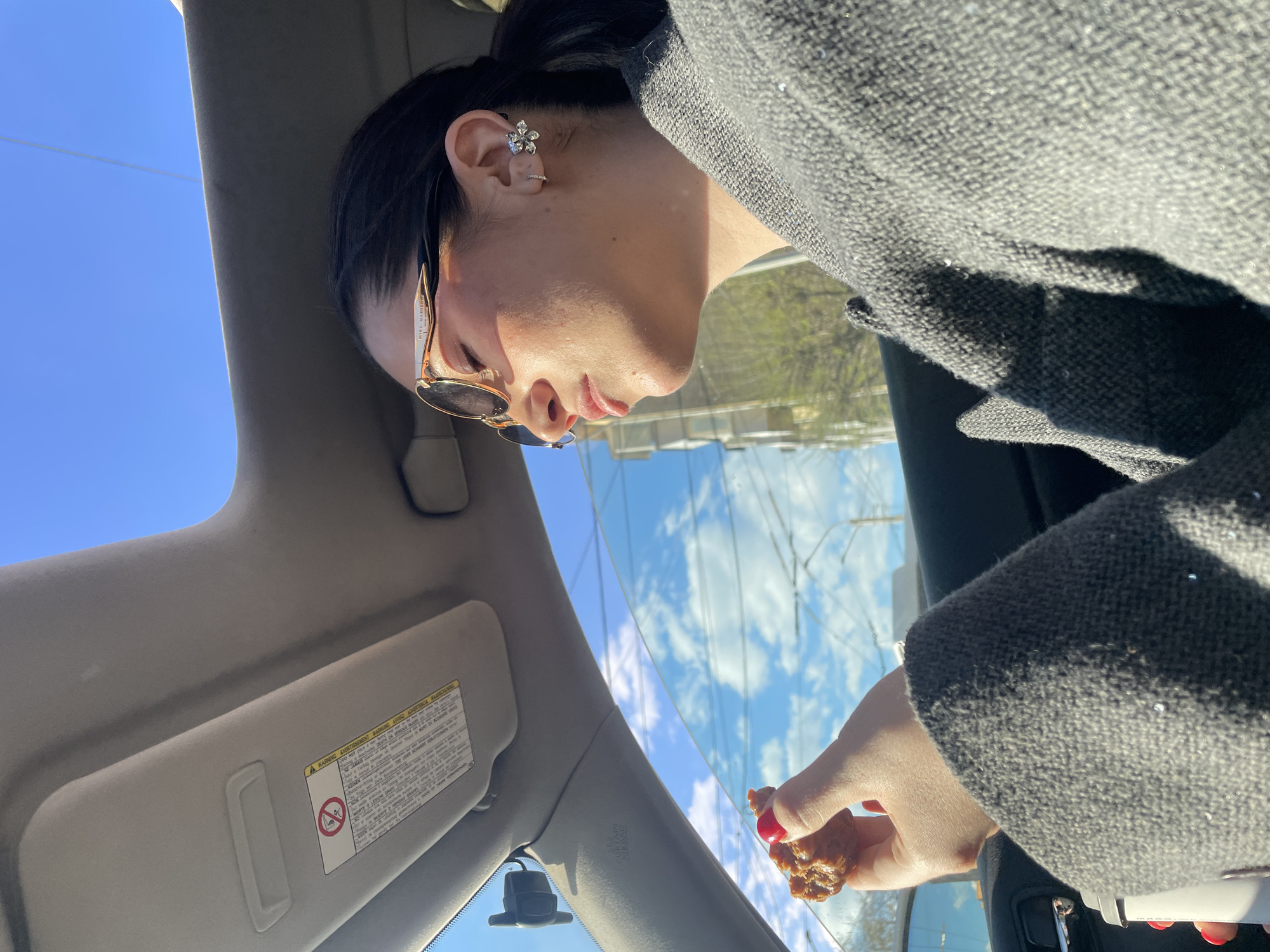 A person wearing sunglasses and a grey wool coat is seated in a car, holding a piece of food. The car's sun visor is visible, as is a rearview mirror. Outside the car window is a building with a glass facade, reflecting the blue sky and clouds. The city of Krasnoyarsk, Russia cannot be verified from the image.