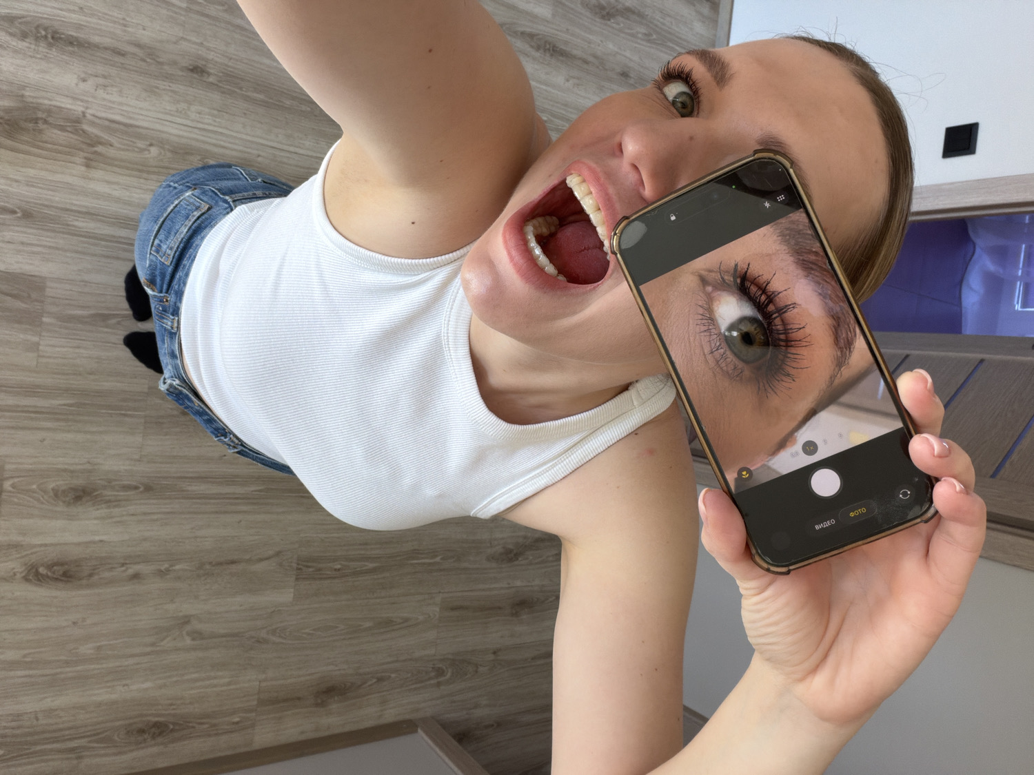 An overhead shot captures a young woman in an indoor setting, likely in Dreiliņi, Latvia. She is positioned on a light wood-look floor, looking upwards and outwards towards the camera with a wide-eyed, open-mouthed expression that conveys surprise or playfulness. Her mouth is agape, revealing her teeth and tongue, and she wears a white ribbed tank top and blue jeans.
In her right hand, she prominently holds a smartphone with a gold-colored frame. The phone's screen displays a striking close-up image of an eye, presumably her own, with long, dark, defined eyelashes, looking directly at the viewer. The screen also shows the interface of a camera application, featuring a large white circular shutter button at the bottom center. Visible text on the screen includes "ФОТО," which means "PHOTO" in Cyrillic (Russian), indicating the current camera mode, alongside zoom options like "1x" and "0.5x."
The background includes a light-colored wall in the upper right, with a black square object, possibly a light switch or outlet, and a darker piece of furniture or fixture below it. The scene appears brightly lit, suggesting it is daytime. The woman seems to be engaged in taking a selfie or a creative self-portrait, using her phone's display to frame her eye.