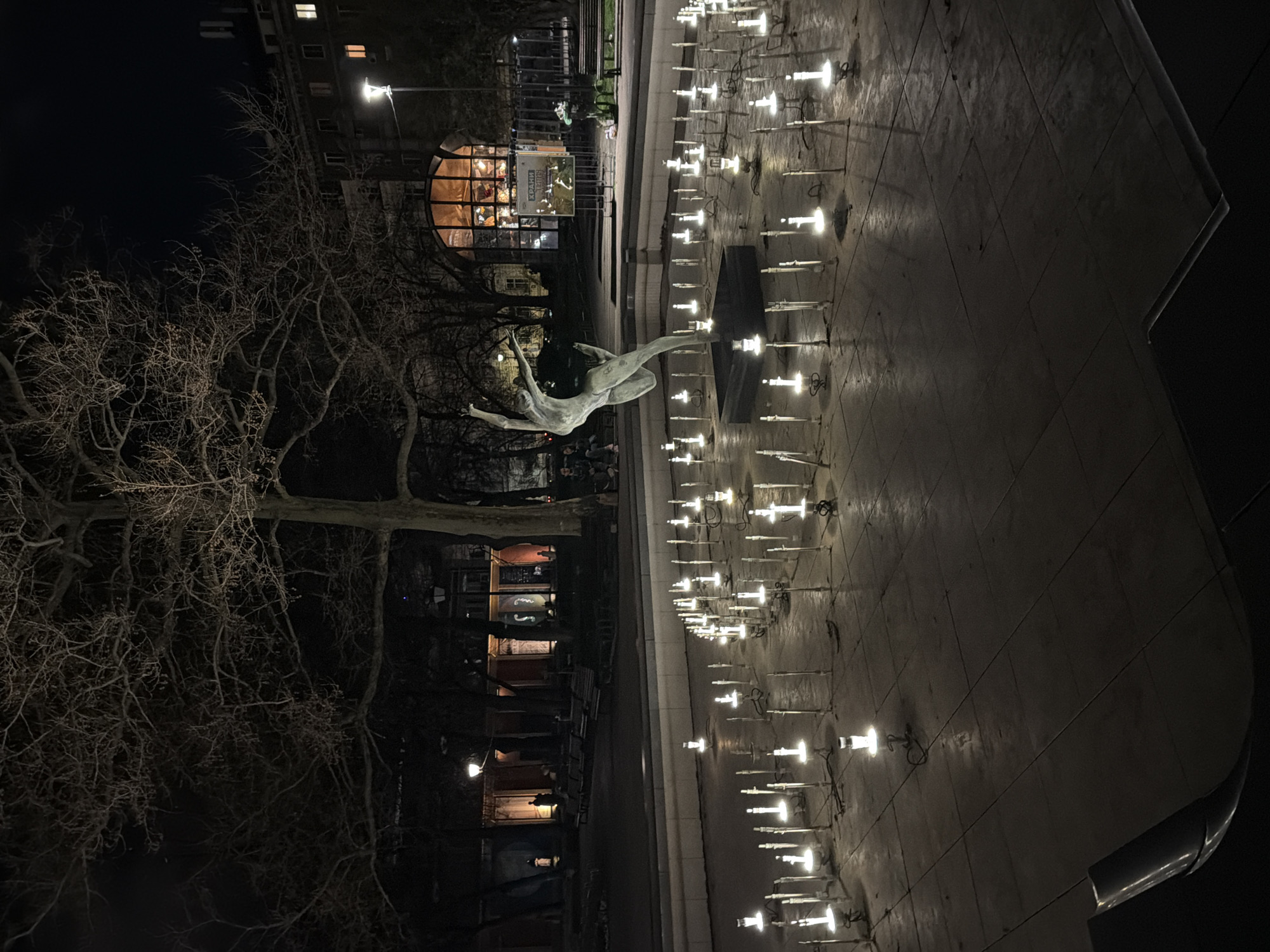 The image shows a night scene in an urban environment. Dominating the frame is a large, outdoor fountain with numerous small lights illuminating the water jets. A bronze sculpture of a human figure, possibly leaping or dancing, is positioned near the center of the fountain. To the left, a large, bare tree is visible, its branches reaching up into the night sky. In the background, several buildings can be seen, including one with a distinctive round window, suggesting a restaurant or a unique architectural feature. The fountain and surrounding area appear to be a public space, possibly a plaza or park within the city. The city of Unknown, Unknown is not mentioned in the image.
