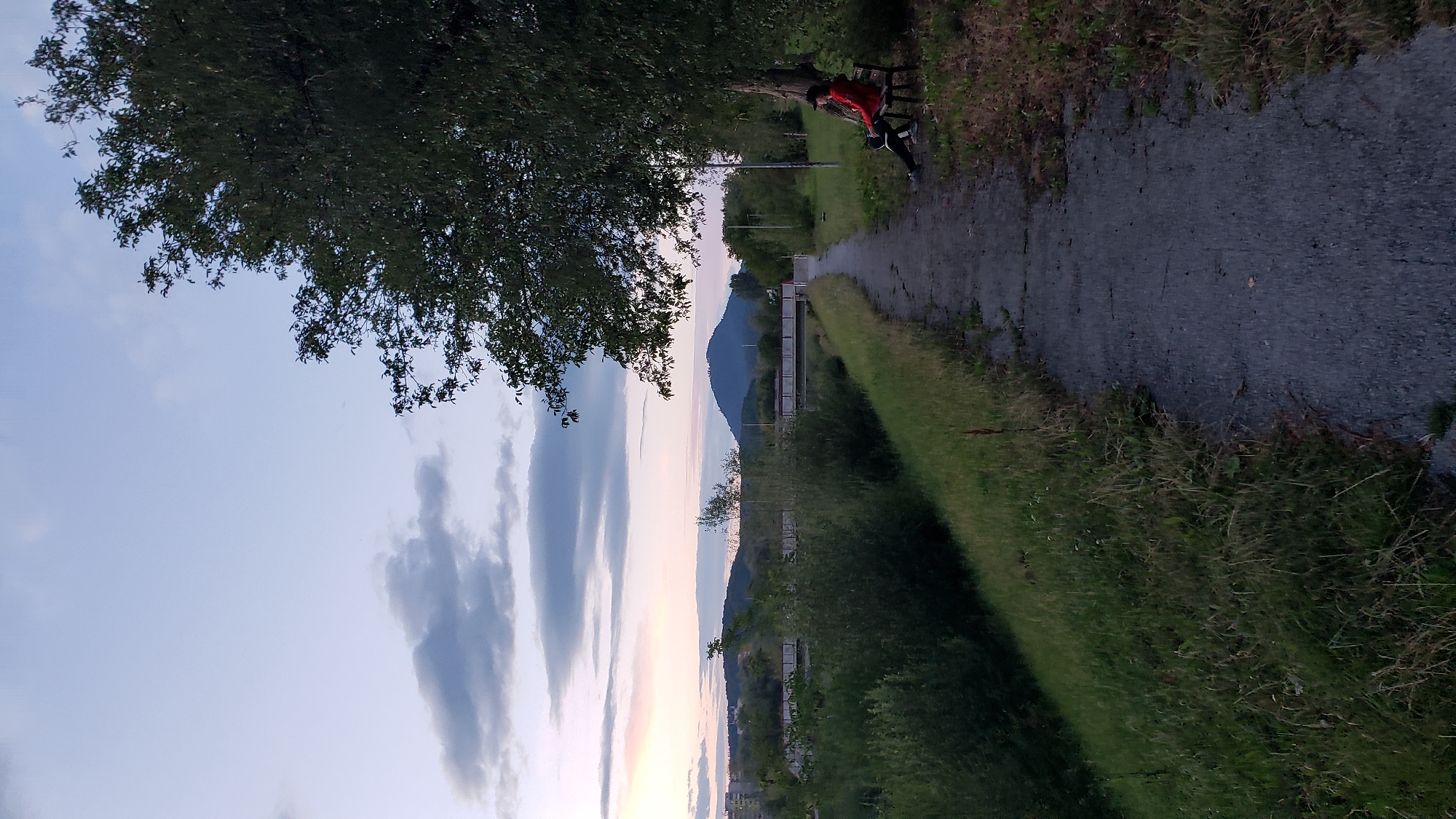 The image depicts a pathway along a grassy embankment, leading towards a bridge spanning a body of water. On the right side of the pathway, a person is seated on a bench under a large tree, wearing a red jacket and dark pants. The landscape includes hills and buildings in the distance, under a cloudy sky with hints of sunset colors. This scene is located along the Torysa river in Prešov, Slovakia.